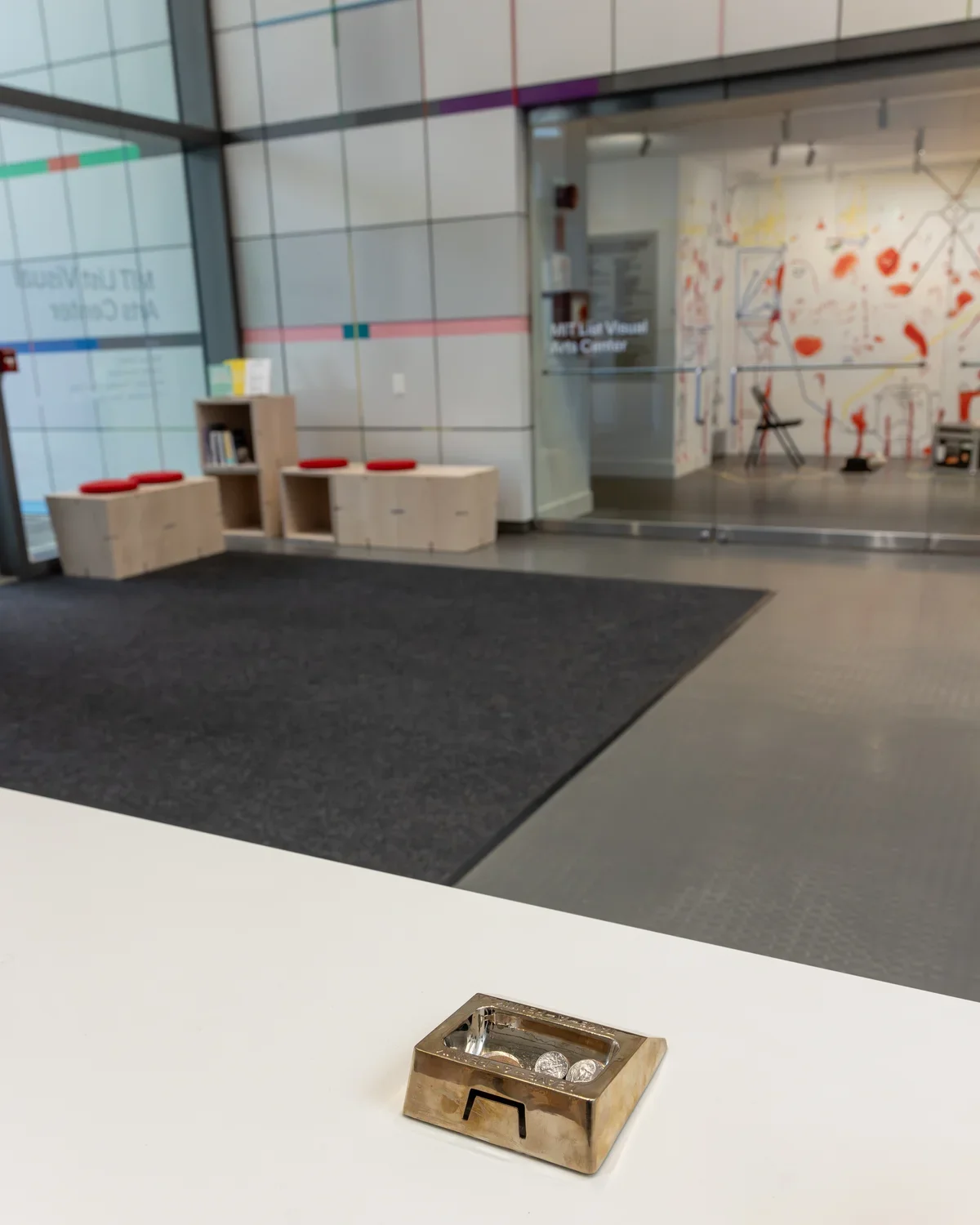 A small brass dish holding a few coins rests on a white counter in the foreground. Behind it, a lobby leads to a translucent glass doors marked "MIT List Visual Arts Center," revealing an exhibition space with white walls covered in red and yellow markings.