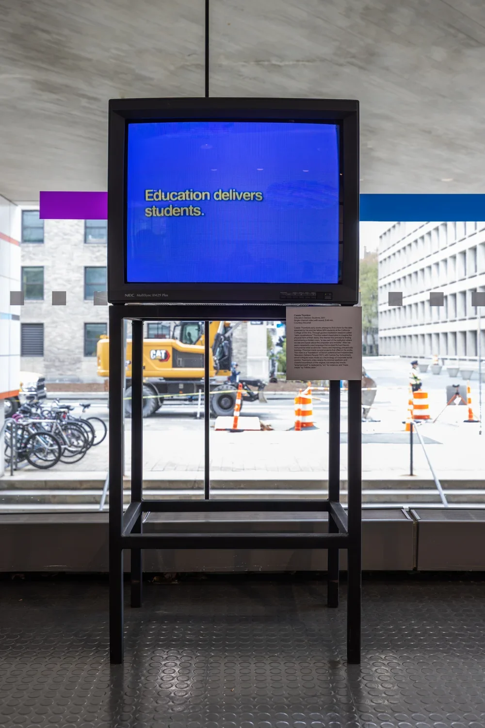 A boxy monitor is positioned against a window, through which a street, construction equipment, and the buildings of a campus are visible. The screen shows white text against a bright blue background, reading, “Education delivers students.”