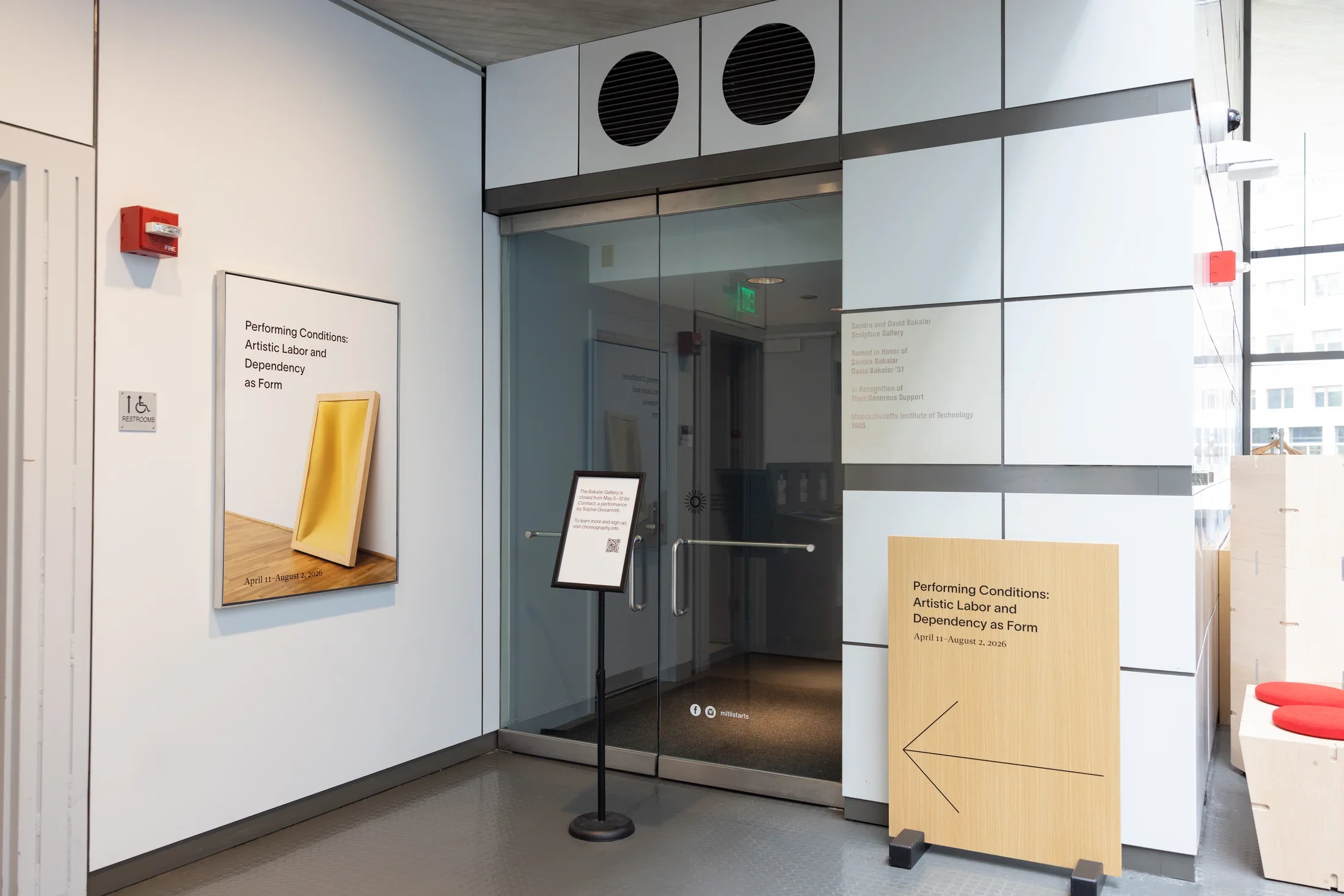 Double glass doors lead from a larger interior space to a short corridor. The doors are blocked by a standing sign with text and a QR code. They are flanked by two larger signs—a poster featuring a rectangular leaning sculpture on the left, a wooden floor sign with a large arrow on the right—that read “Performing Conditions: Artistic Labor and Dependency as Form.”