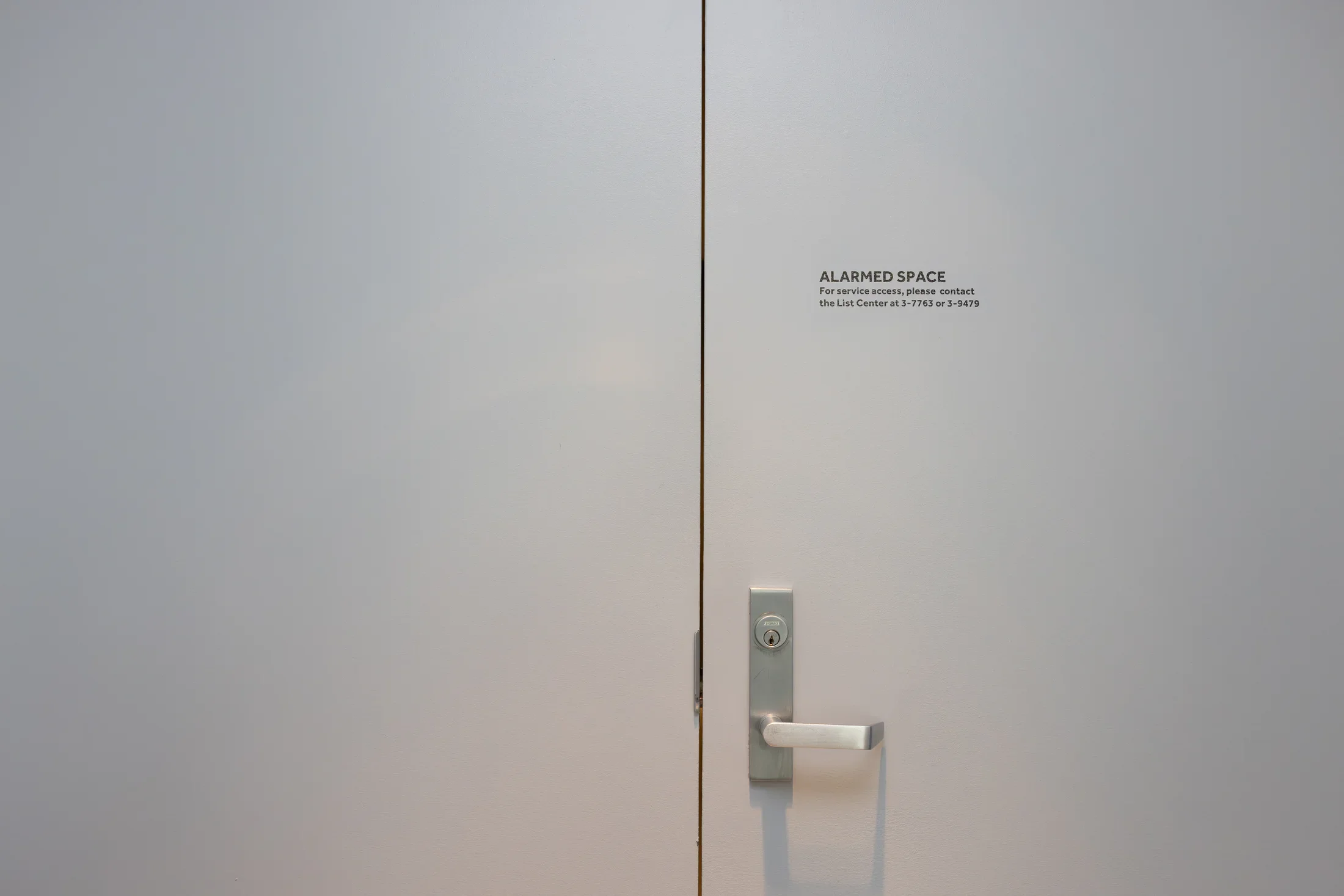 The entire frame is occupied by the flat, gray, nearly blank expanse of closed double doors, split down the middle by the edge where they meet. A metal door handle and keyhole is visible on the right door towards the bottom; towards the top, small text printed on the door reads, “ALARMED SPACE / For service, please contact the List Center at 3-7763 or 3-9479.”