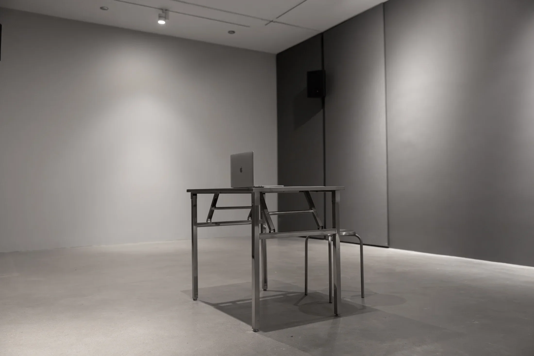 Two pieces of spare, angular, metal furniture occupy an otherwise empty space with gray walls and a concrete floor: a small table and a stool. An open Mac laptop sits on top of the table.