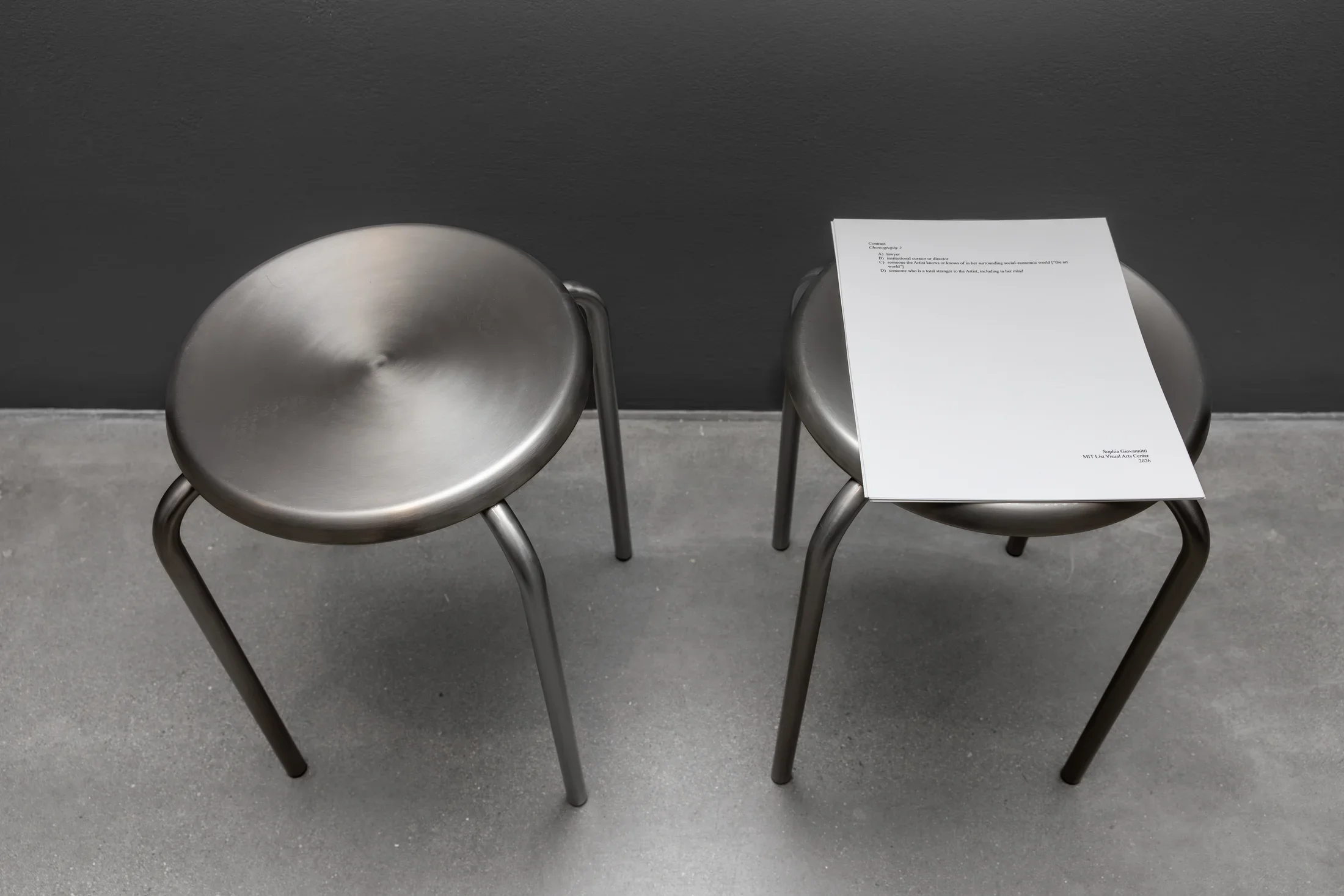 Two identical metal stools sitting side by side are photographed from above. A short stack of paper sits on top of the right stool; it is mostly blank and features a A-D list of possible participants (e.g. “lawyer,” “institutional curator or director”) underneath a title reading “Contract: Choreography 2.”