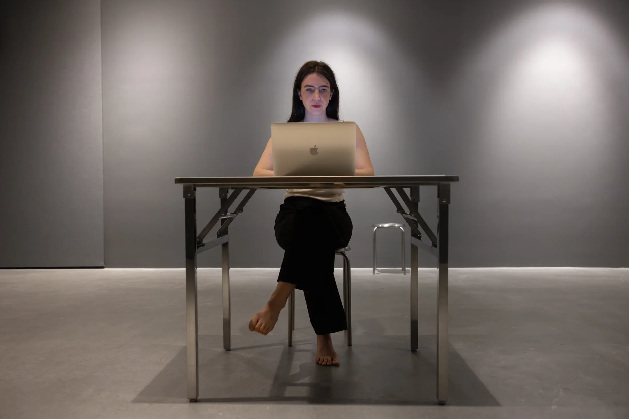 Austere metal furniture—a small table and two stools—occupy an otherwise empty space with gray walls and a concrete floor. A woman sits behind the table on one of the stools, facing the camera and working on a laptop. Her feet are bare.