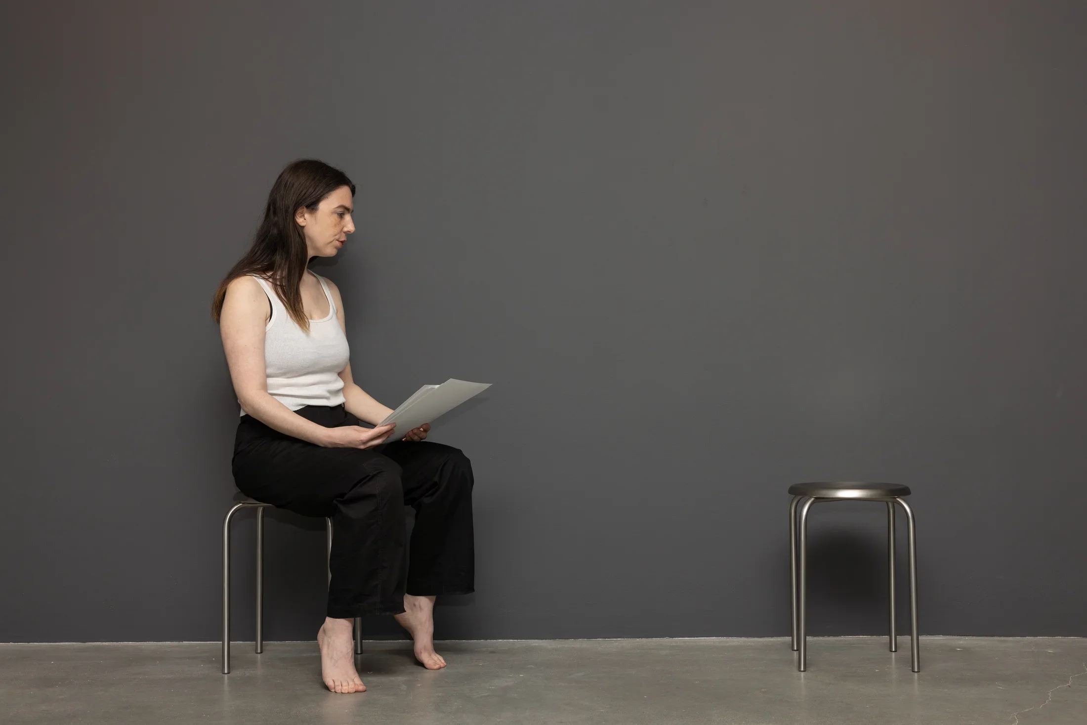 Two metal stools sit against a dark gray wall. A woman with a white tank top, black pants, and bare feet sits on the left stool and faces the unoccupied one. She holds some papers in her hands.