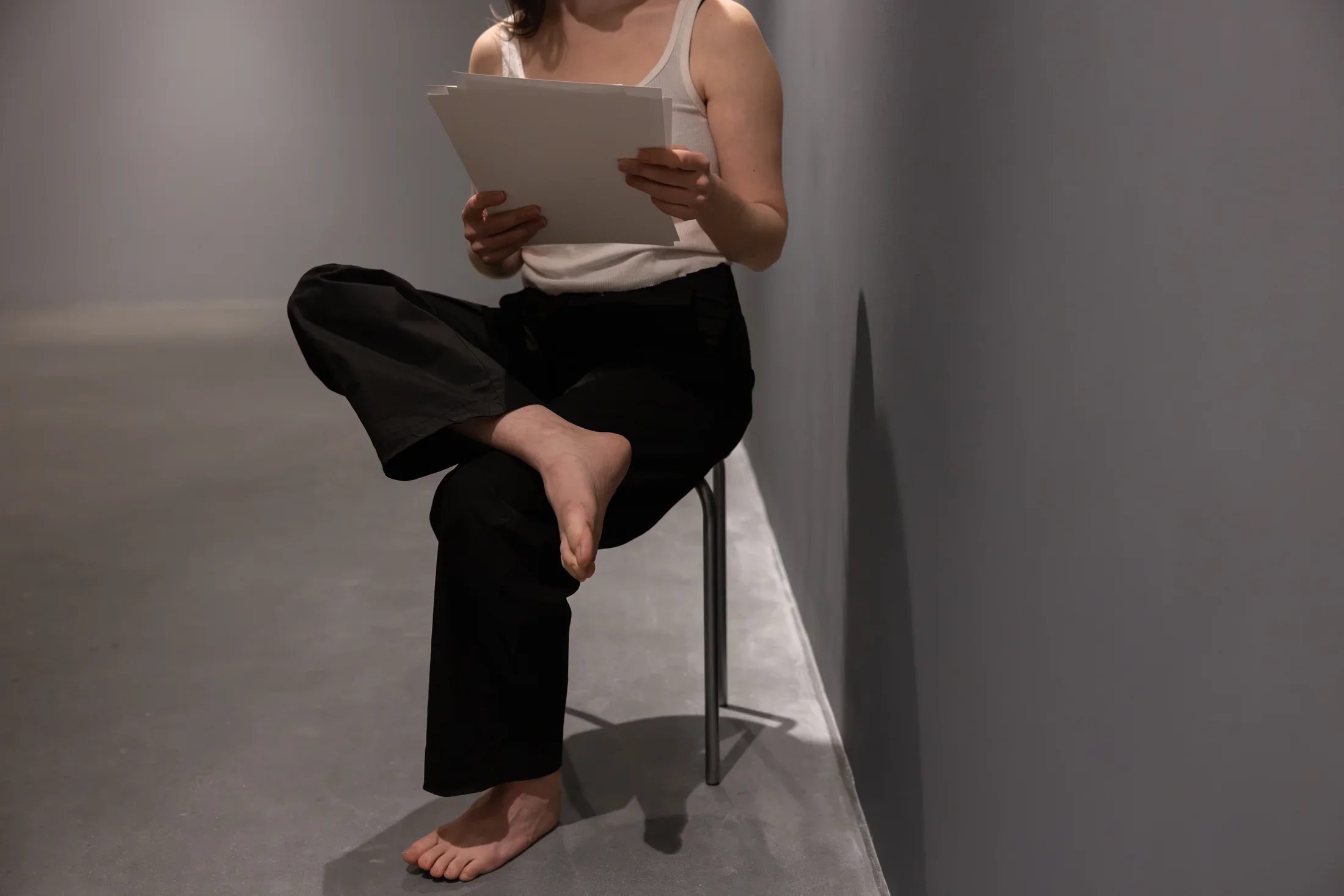 A woman wearing a white tank top and black pants sits on a metal stool, crossing her legs and holding some papers in her hands. Her body faces the camera, but the photograph’s framing crops out her face.
