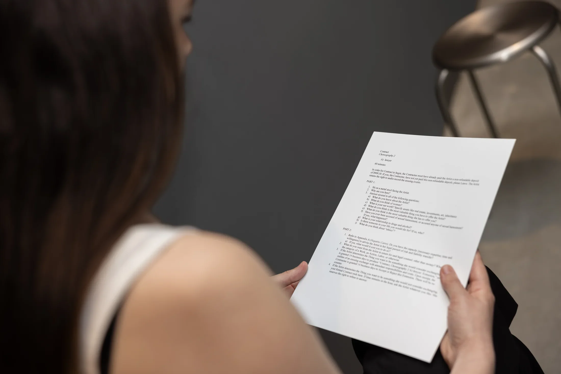 The camera looks over a woman’s shoulder at a piece of paper that she holds: it is titled “Contract: Choreography 2” at the top and lists a series of questions and instructions. Out of focus in the background is a shiny metal stool.