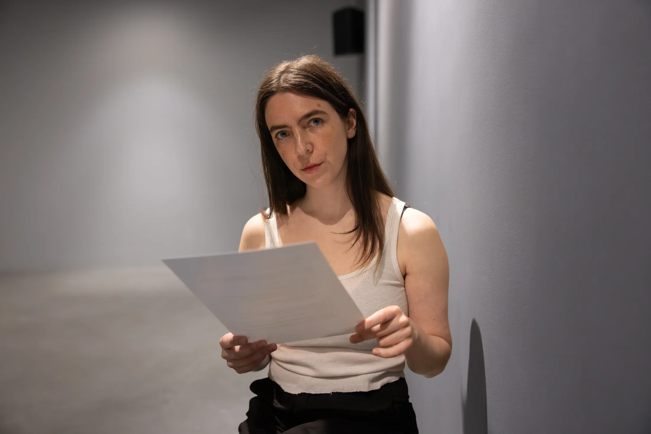 A woman wearing a white tank top and black pants makes eye contact with the camera and holds some papers in her hands. She appears to occupy a bare space with gray floor and walls.