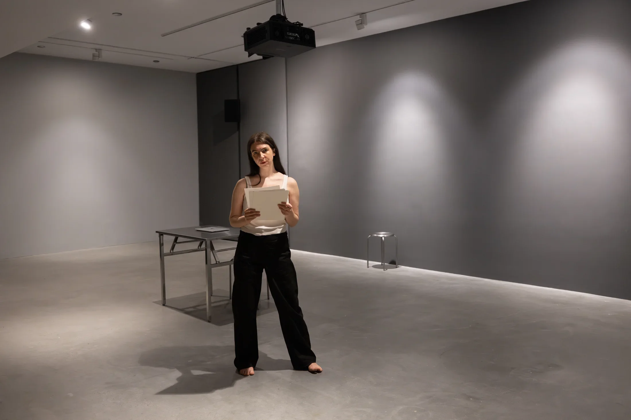 A woman wearing a white tank top and black pants stands in a gallery, looking towards the camera while holding some papers. Metal furniture—a small table and stool—is visible behind her in an otherwise austere space with gray walls and floor.