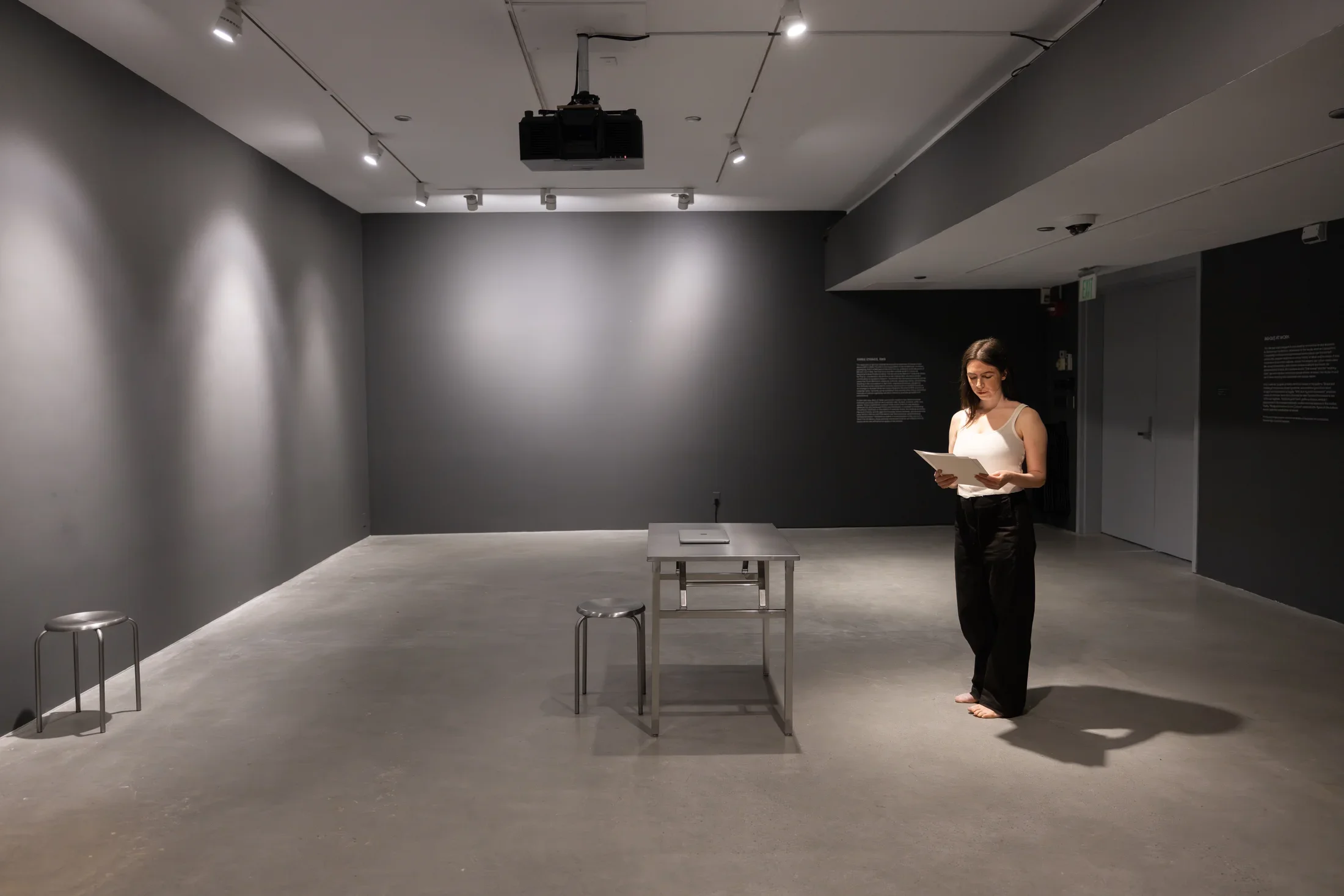 A woman wearing a white tank top and black pants stands in a gallery, looking down at some papers she holds. Metal furniture—two stools and small table with a closed laptop on it—are the only other objects in the austere, gray-painted space.