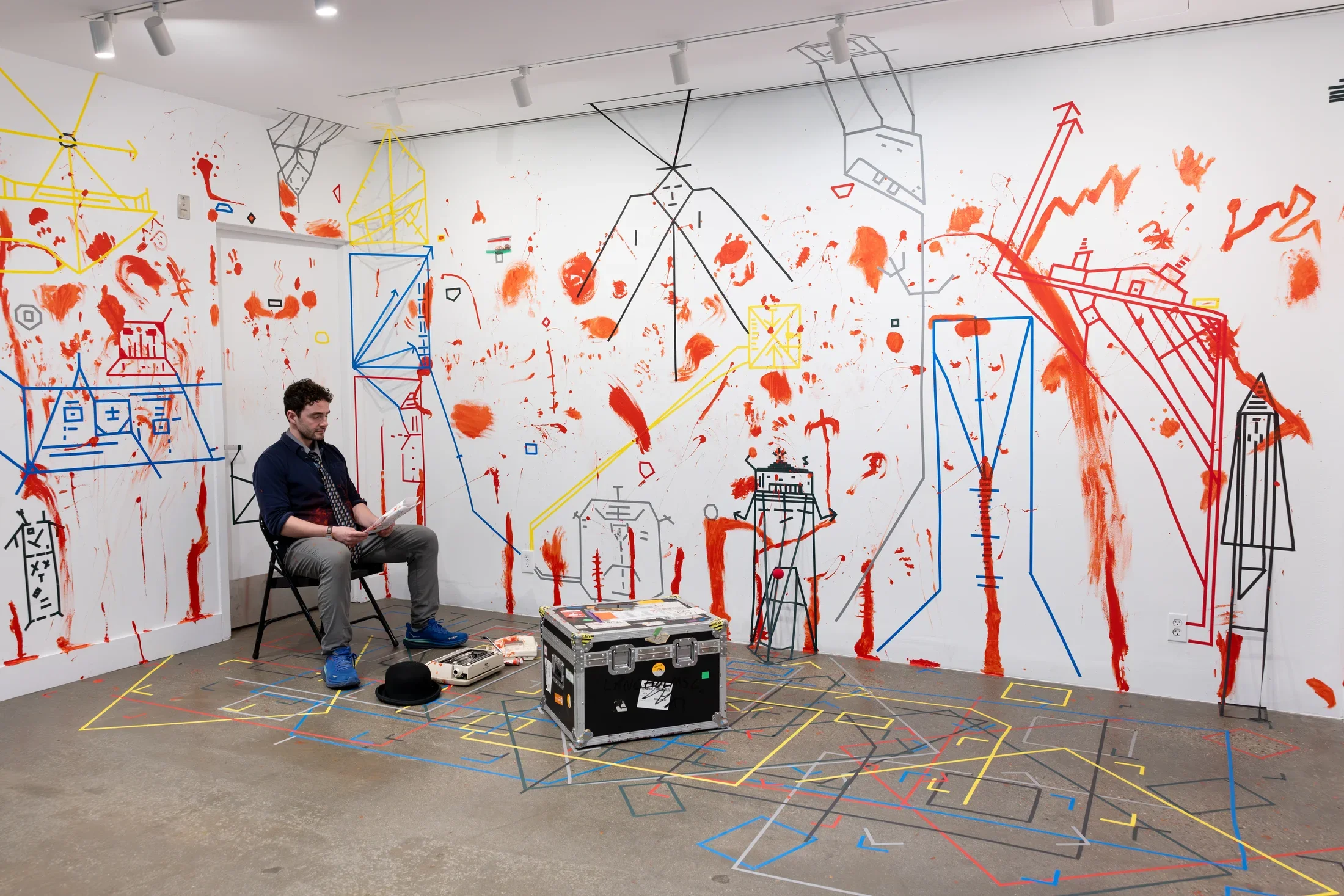 A man sits on a sturdy chest or box and looks down at the packet of paper in his hands. The white walls behind him are marked with dense lines of colored tape and splotches and smears of red paint. Other objects, like a hat and a typewriter, are arranged at his feet.