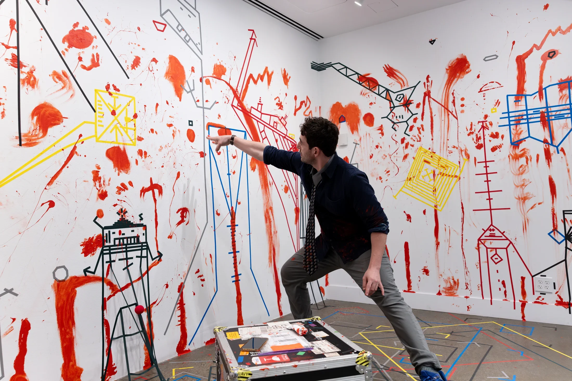 A man wearing a tie stands in a lunge position, one hand pointing at a splotch of red paint on a white wall. The rest of the wall is covered with dense lines of colored tape and splotches and smears of paint.