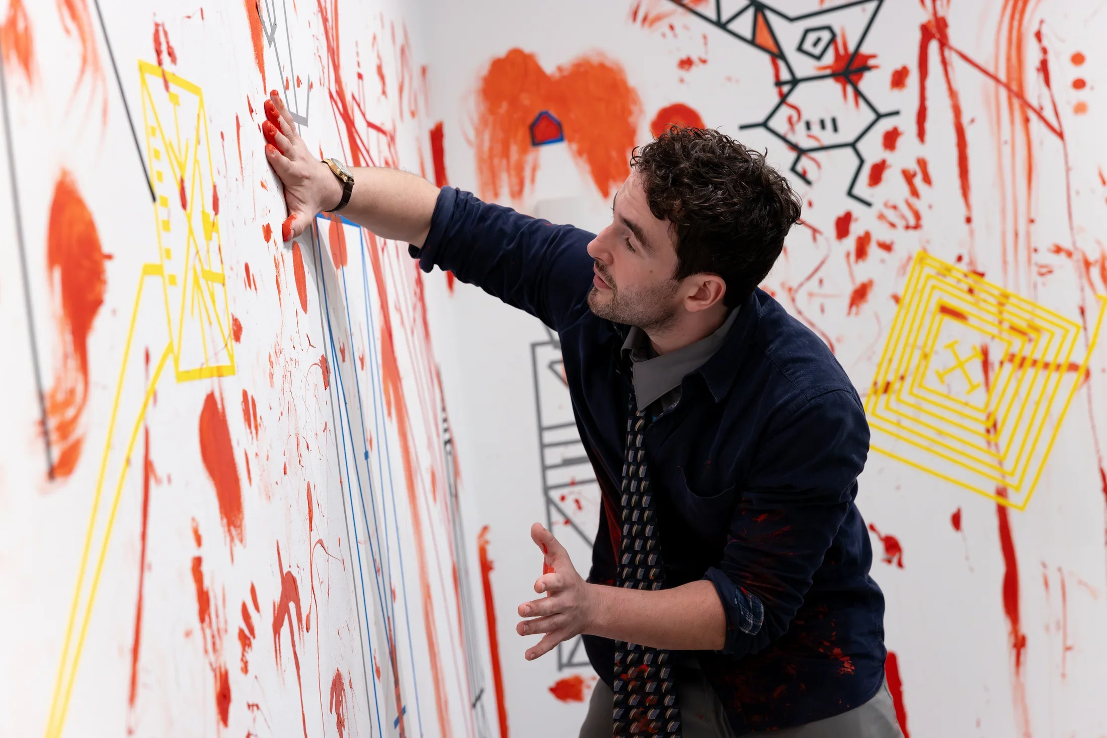 The top half of a man wearing a collared shirt and tie is shown against the background of white walls covered in tape diagrams and irregular red paint. He uses one palm to smear or apply paint onto the wall in front of him.