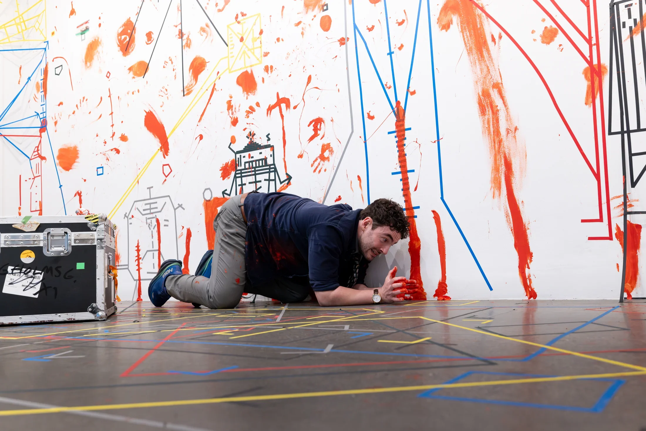 A man rests on his elbows and knees in a gallery, his hands covered in red paint. The white walls behind him are covered in geometric forms made of colored tape, and handprints and smears of red paint. The gray floor is also marked with crisscrossing lines of tape.