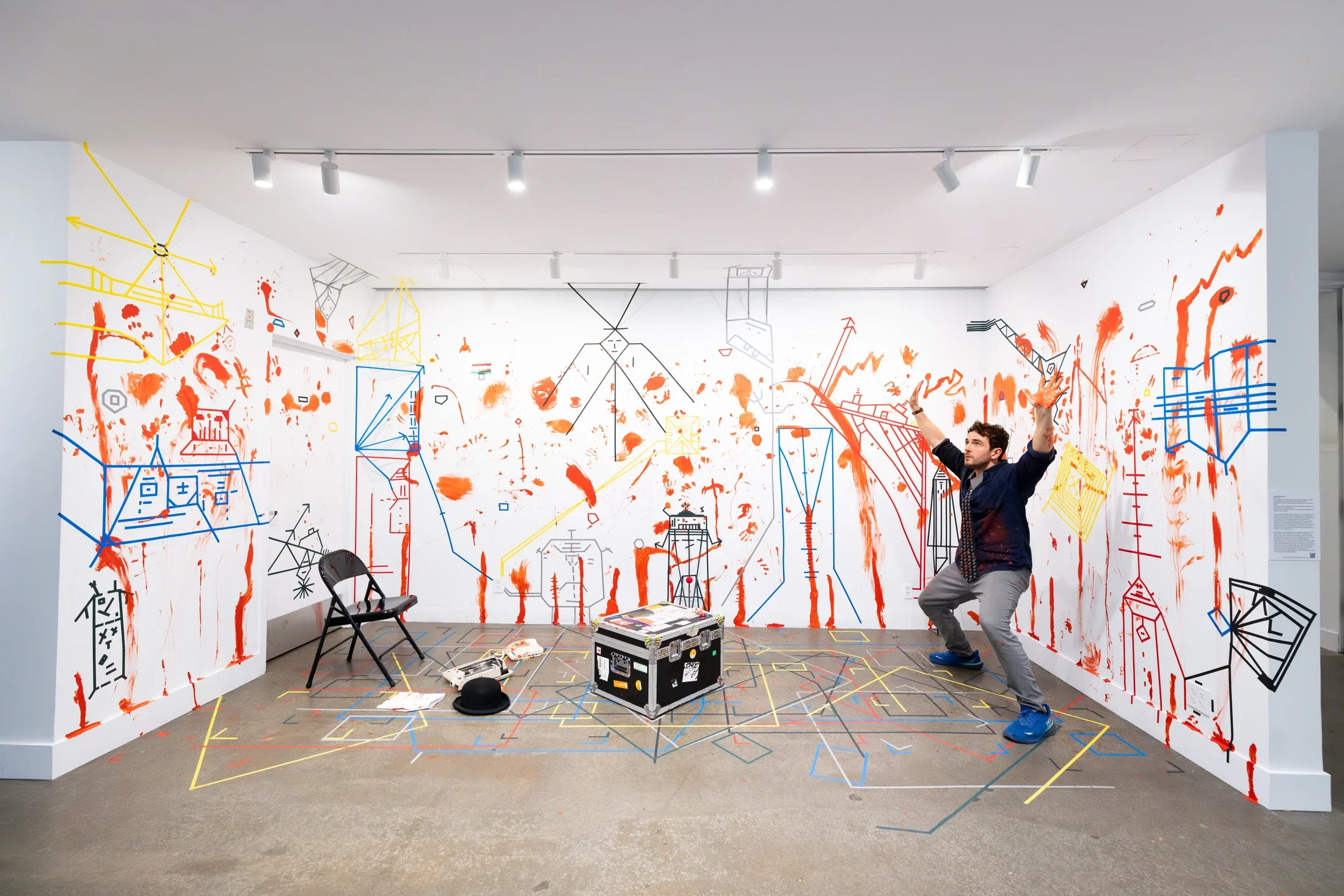 A man wearing a tie stands in a theatrical pose in a gallery: knees slightly bent, arms above his head, palms upturned. The walls and floor around him are marked with dense lines of colored tape and splotches and smears of red paint. A few props, like a hat, a typewriter, and a phone, sit on the floor too.
