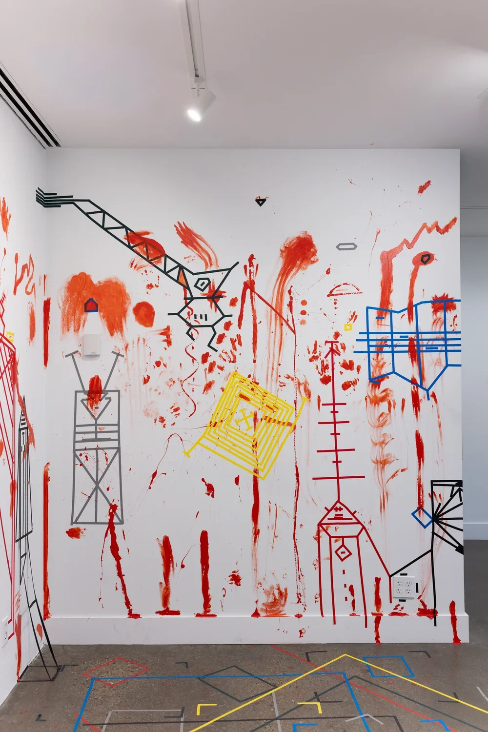 A white wall—densely marked with handprints and vertical trails of red paint, as well as obscure geometric figures made of colored tape—occupies most of the frame. The portion of the gray floor that is visible is also marked with boxes and brackets of multicolored tape.