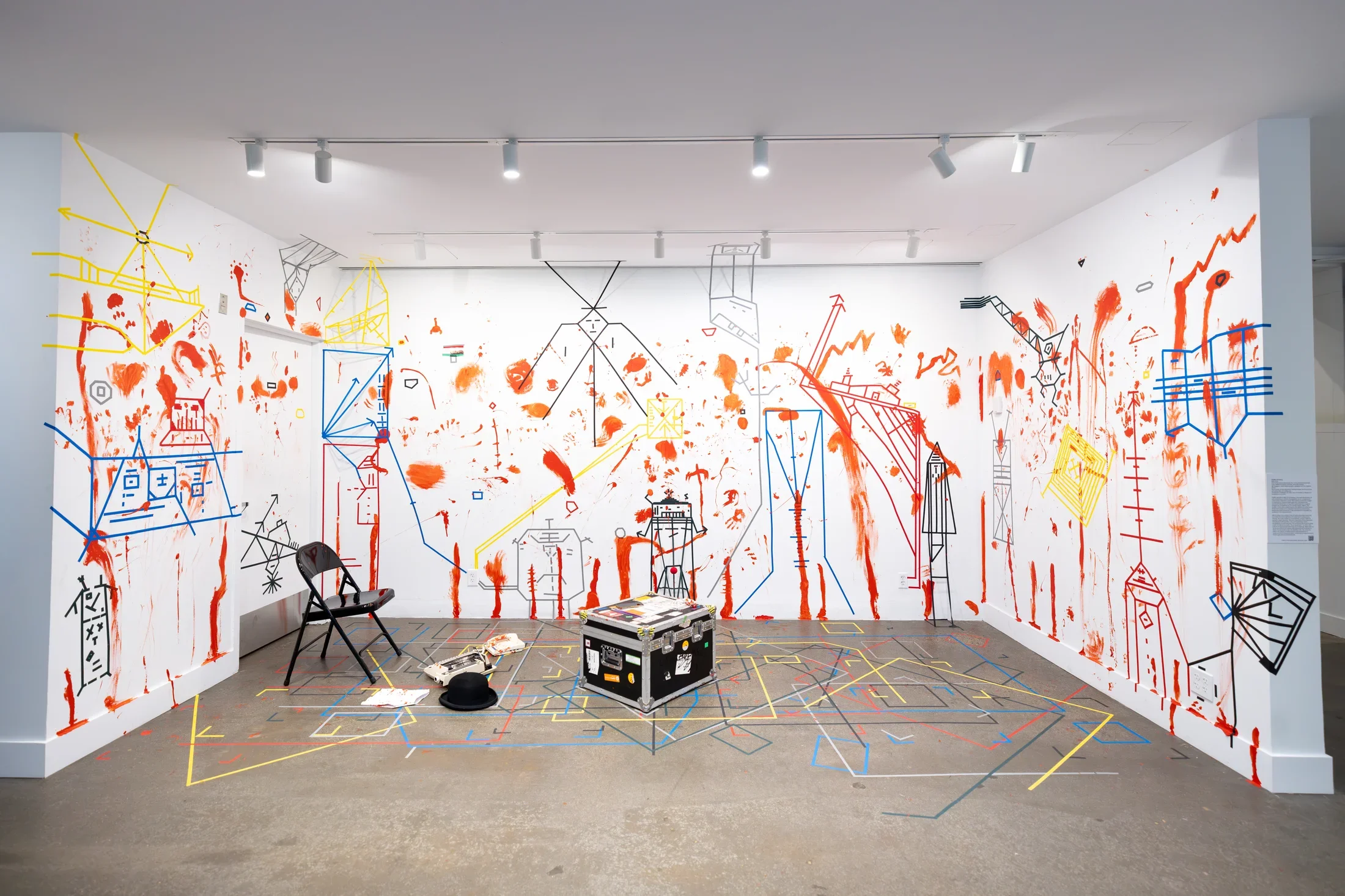 Three walls and the floor of a gallery are densely marked—with handprints and vertical smears of red paint, and with geometric figures and boxes made of colored tape. A few objects, including a metal folding chair, a shipping chest, a typewriter, and a phone, sit on the floor atop the tape markings.