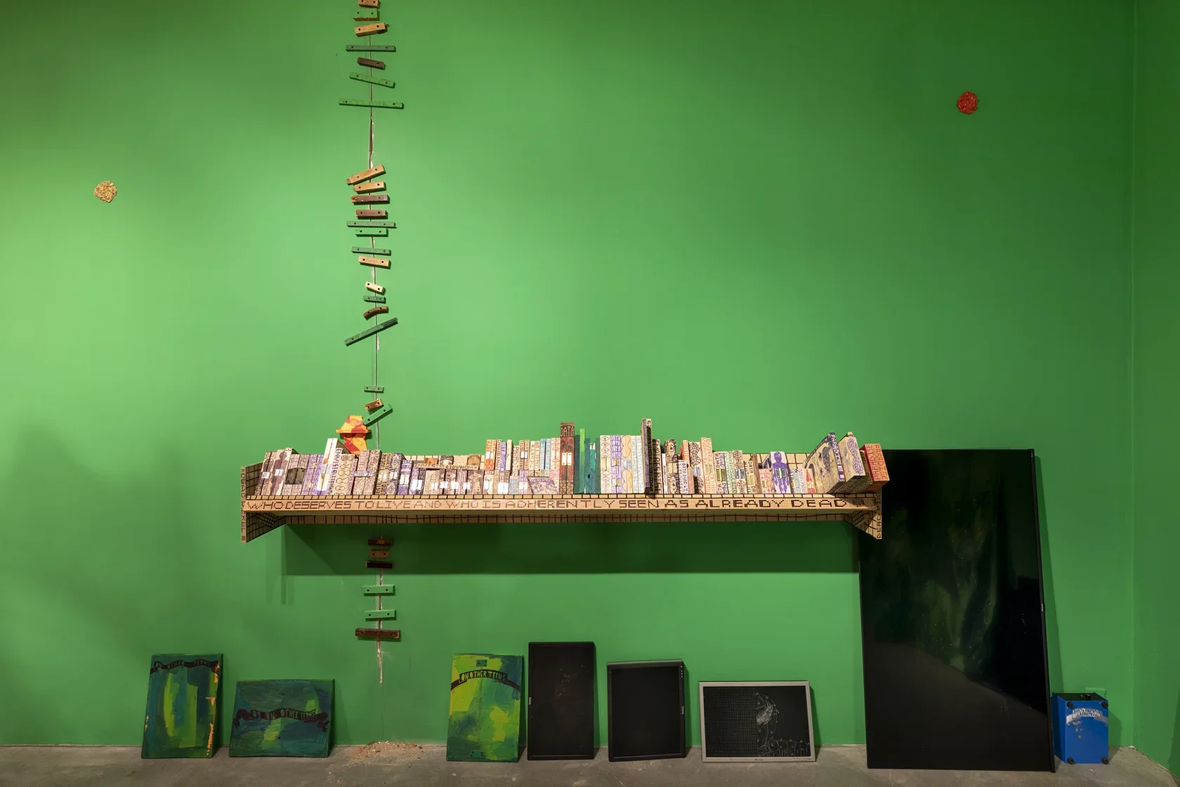 A bright green wall displays a long shelf filled with small, colorful, rectangular wooden objects, some with visible text. Below the shelf, three green paintings with text, four digital screens, and a blue container are arranged against the wall. To the left, a vertical line of small, colorful wooden blocks extends upwards connecting a seam in the wall.