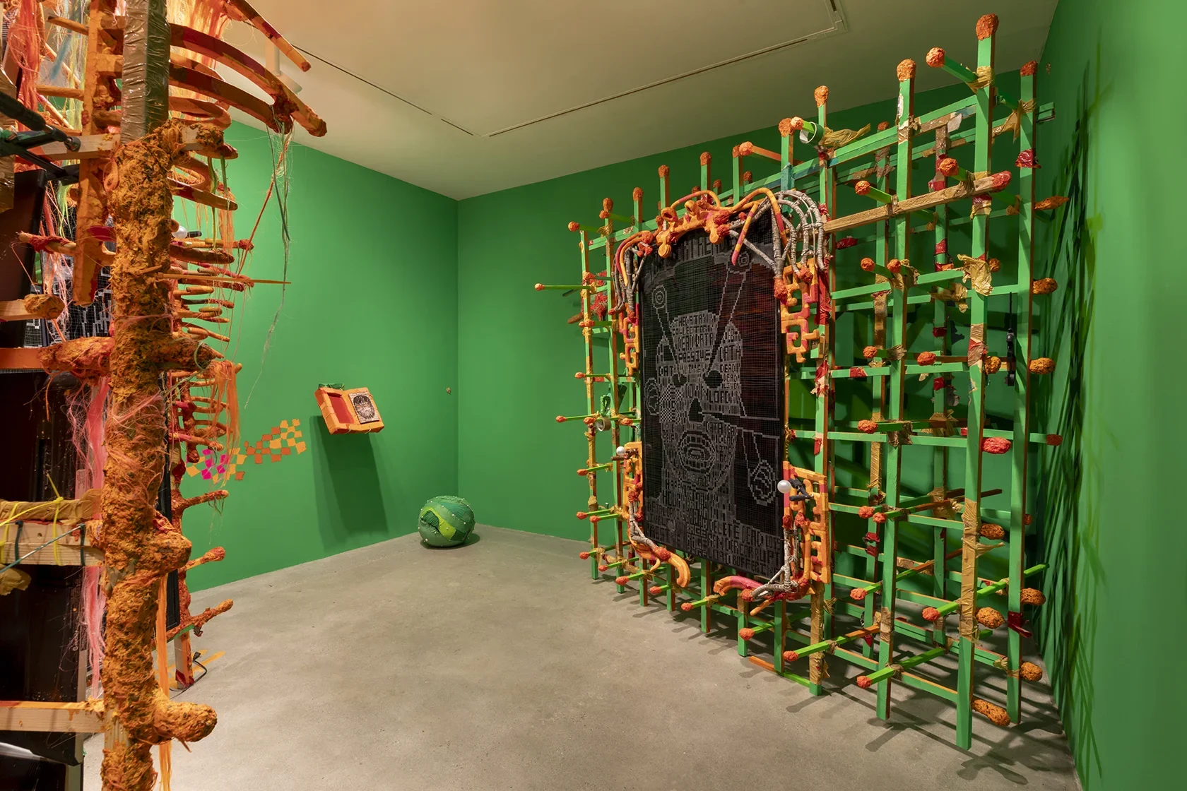 A room with bright green walls and a concrete floor. On the left, a tall, irregular structure of orange and brown textured materials stands. On the right, a large rectangular grid of green and orange rods frames a digital screen with white line drawings.