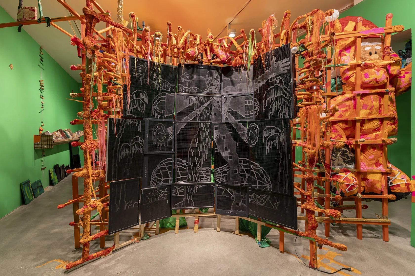 A large art installation features a structure made of wooden slats wrapped with orange tape and yarn in the center of the room. A gridded wall of digital screens with white chalk-like drawings are integrated into the structure, surrounded by various objects including an orange figure in the form of a Santa Claus inflatable.