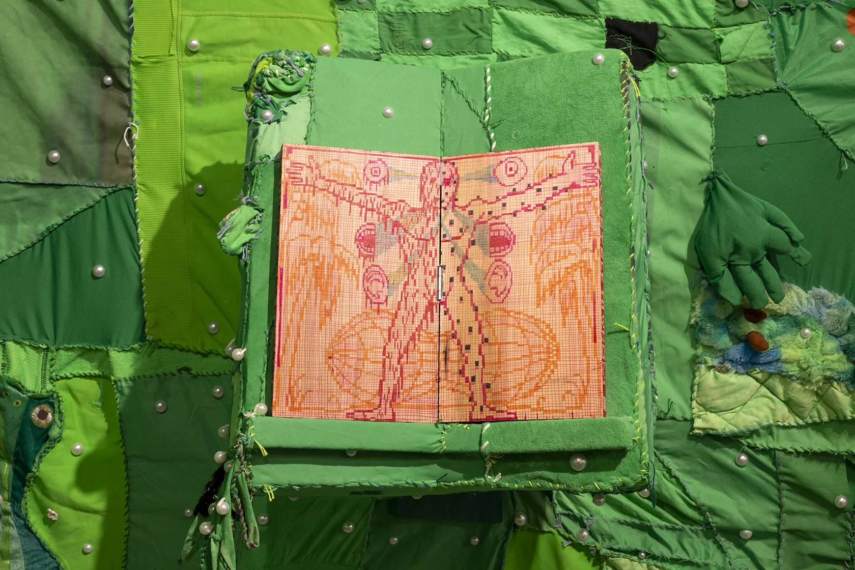 An open wooden book with a pixelated illustration of a human figure in orange, pink, and red is centered on a raised section of green fabric. The fabric is part of a larger green patchwork adorned with white pearls, and a green fabric hand emerges from the right, in front of a patch of colorful textured material.