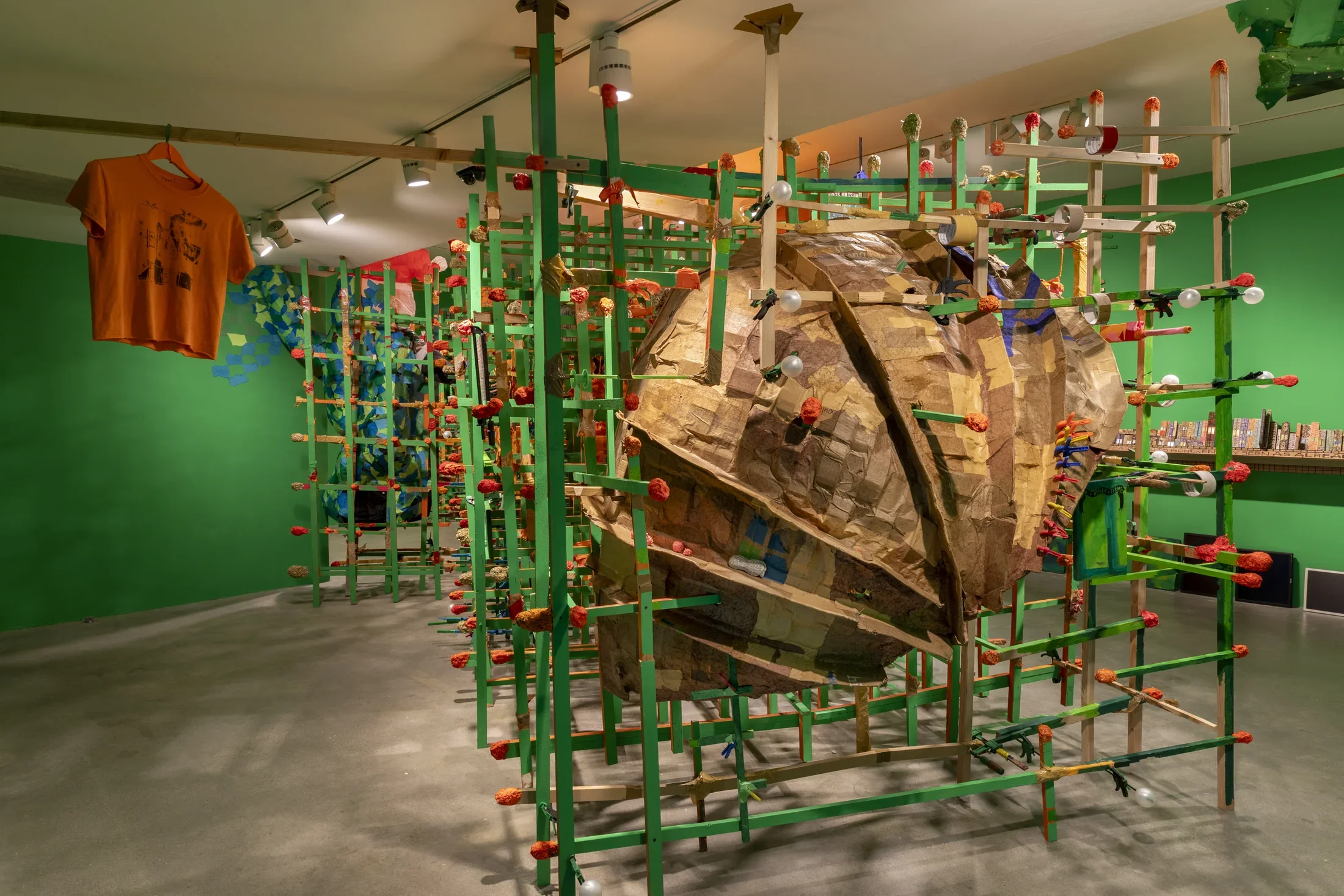 A large, textured, brown cardboard sculpture, is encased within a green wooden grid structure. The installation is set in a room with bright green walls, and an orange t-shirt hangs from the ceiling on the left side of the sculpture.