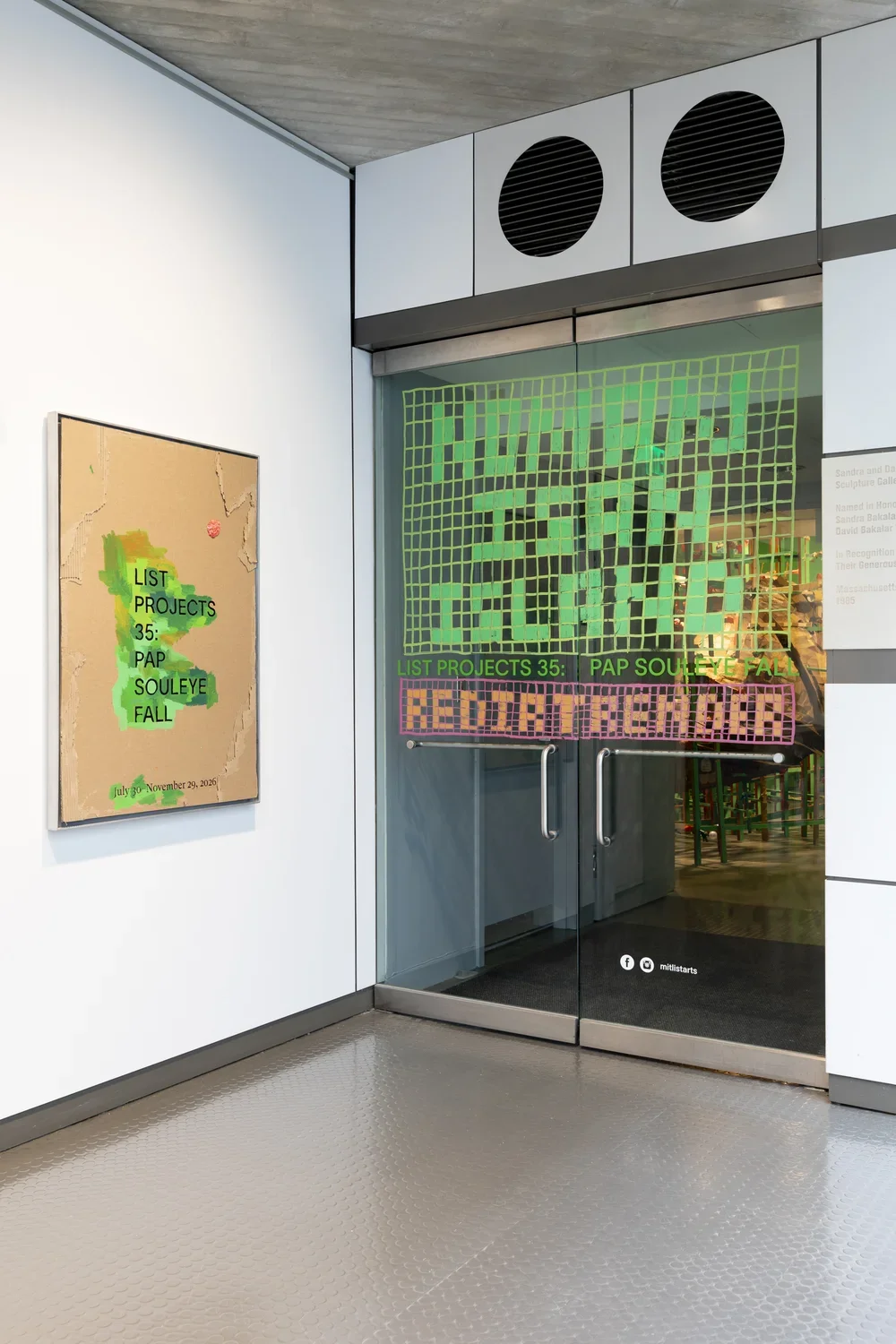 An entrance to a gallery features a cardboard sign on the left wall with green paint surrounding text reading 'LIST PROJECTS 35: PAP SOULEYE FALL' and dates. The glass door on the right has green grid-like text that reads ‘NO MAN IS AN ISLAND’ with orange text below reading ‘REDIRTRENDER’ above the door handles.