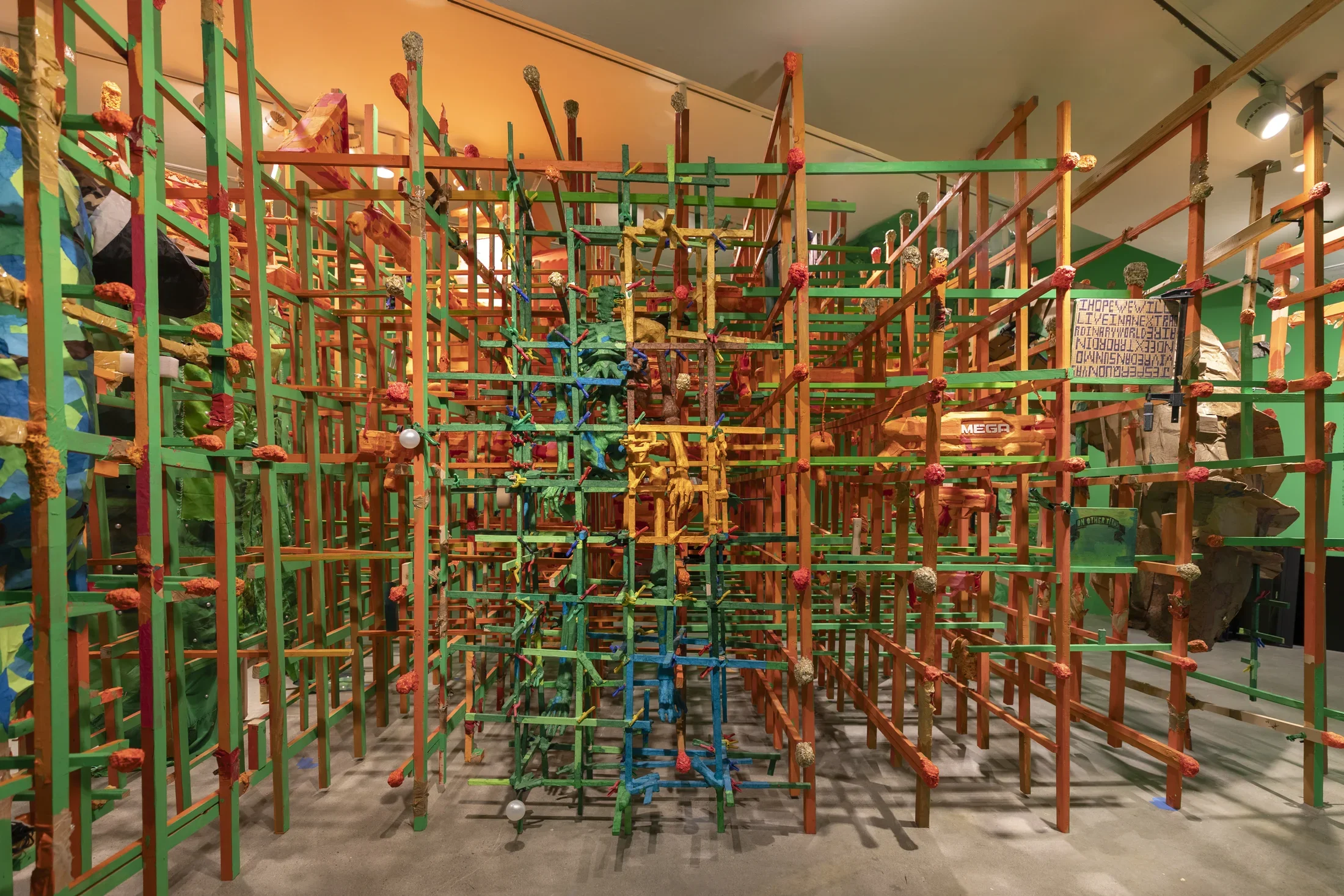 A large, intricate sculpture made of interconnected wooden sticks in green, orange, and blue. The dense, grid-like structure fills the frame, with small red and silver spheres attached at various points. Fabric elements are woven into parts of the central structure.