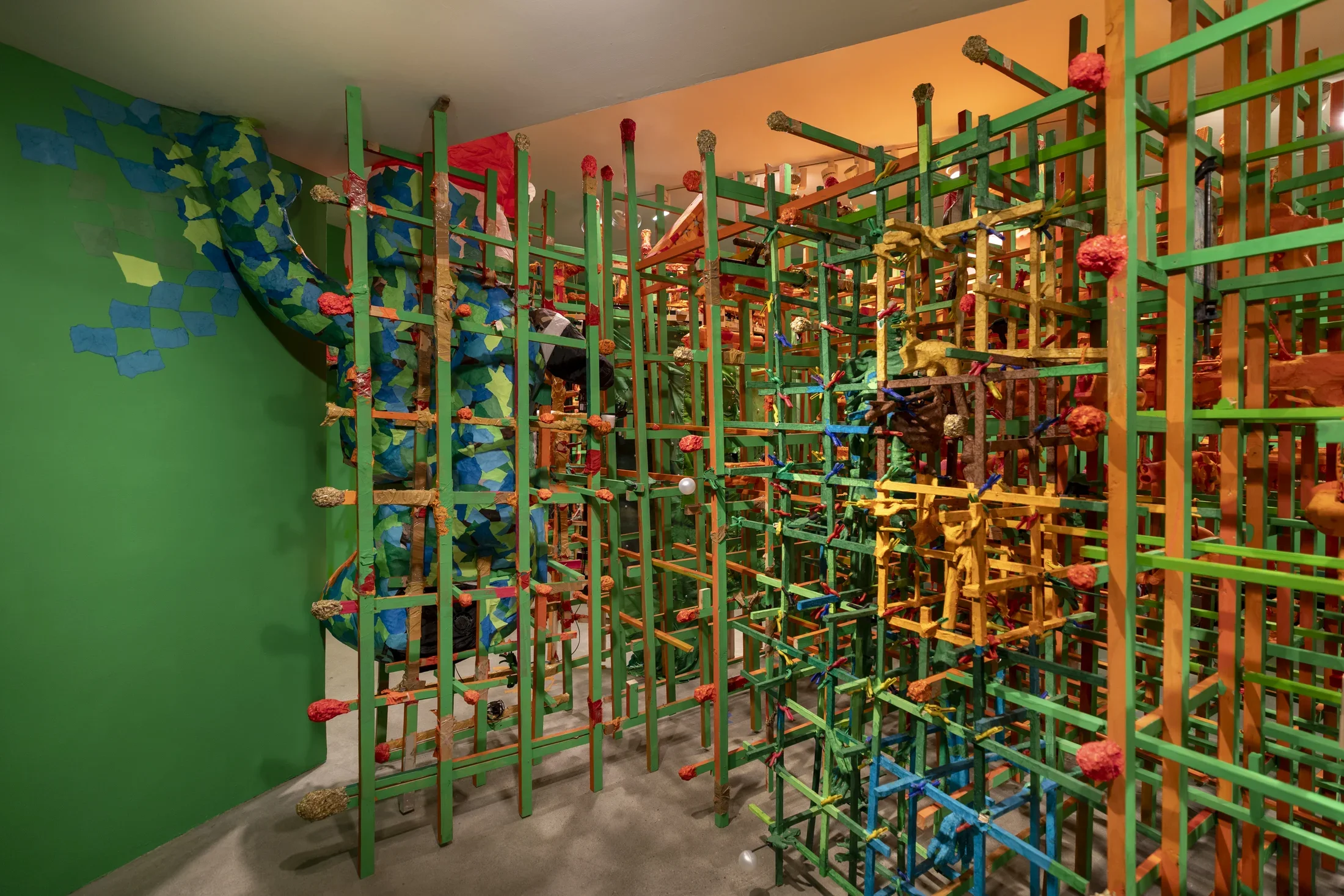 A vibrant, room-filling sculpture features a dense, grid-like structure of green, orange, and blue painted sticks with red spherical elements. On the left, a large, organic form covered in blue and green tessellated pieces emerges from a green wall and intertwines with the stick structure.
