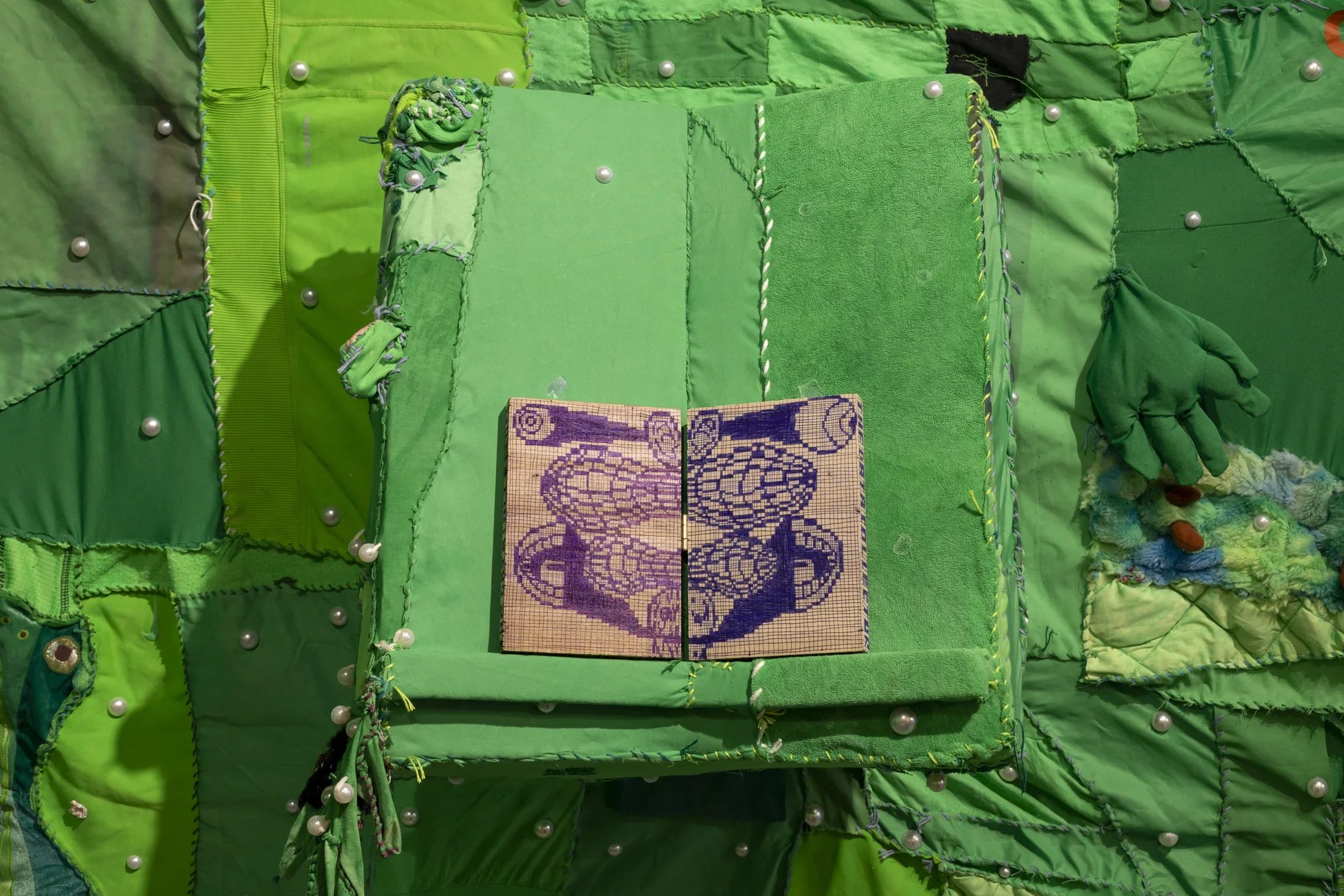 A large textile artwork made of various shades of green fabric patches sewn together, with white pearls embedded throughout. In the center, an open book with a light brown cover and purple and blue patterned pages rests on a raised green fabric. A green glove and other textured fabric elements are sewn onto the right side.