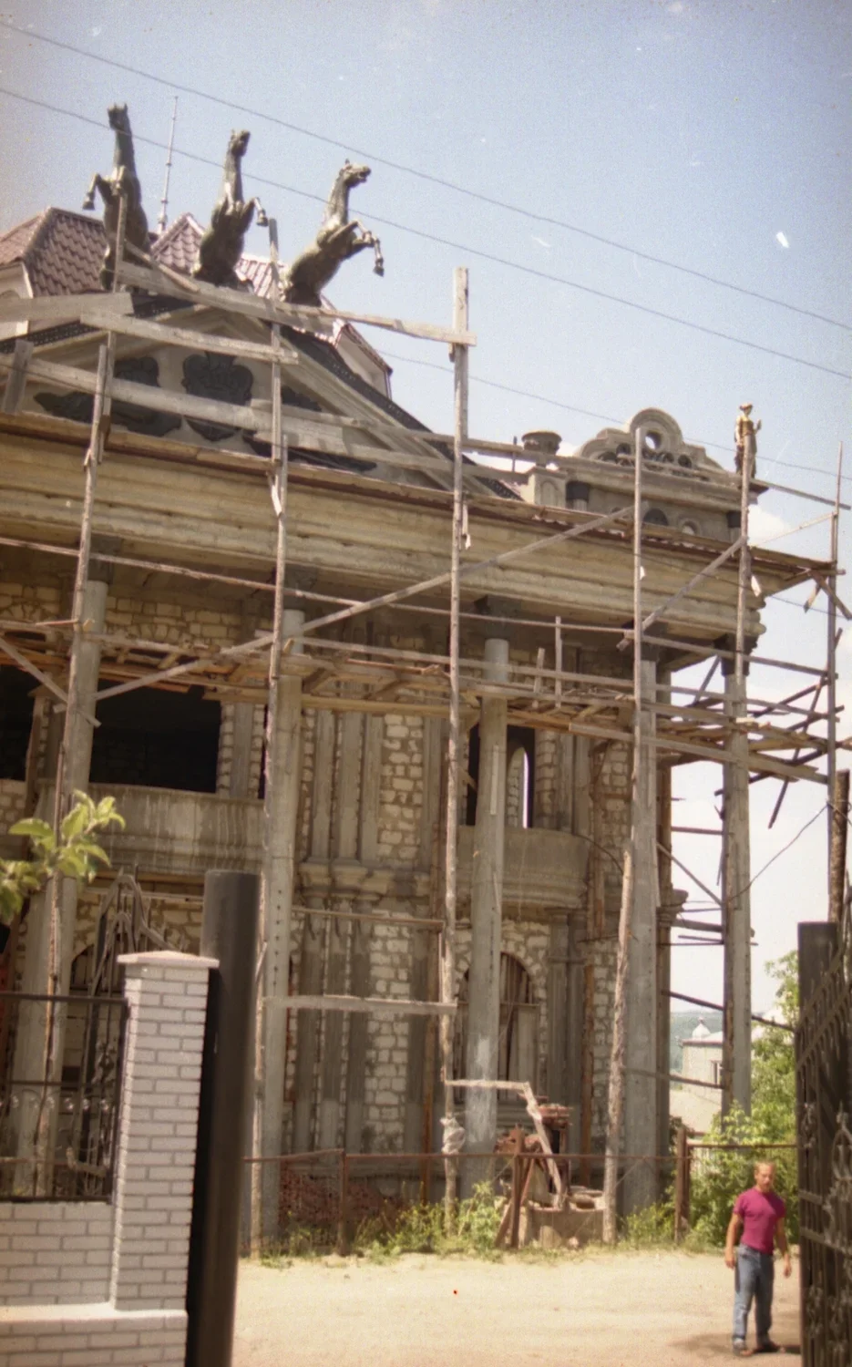 Video still showing a building with wooden scaffolding in front, three horse sculptures on the roof, a man standing in front.