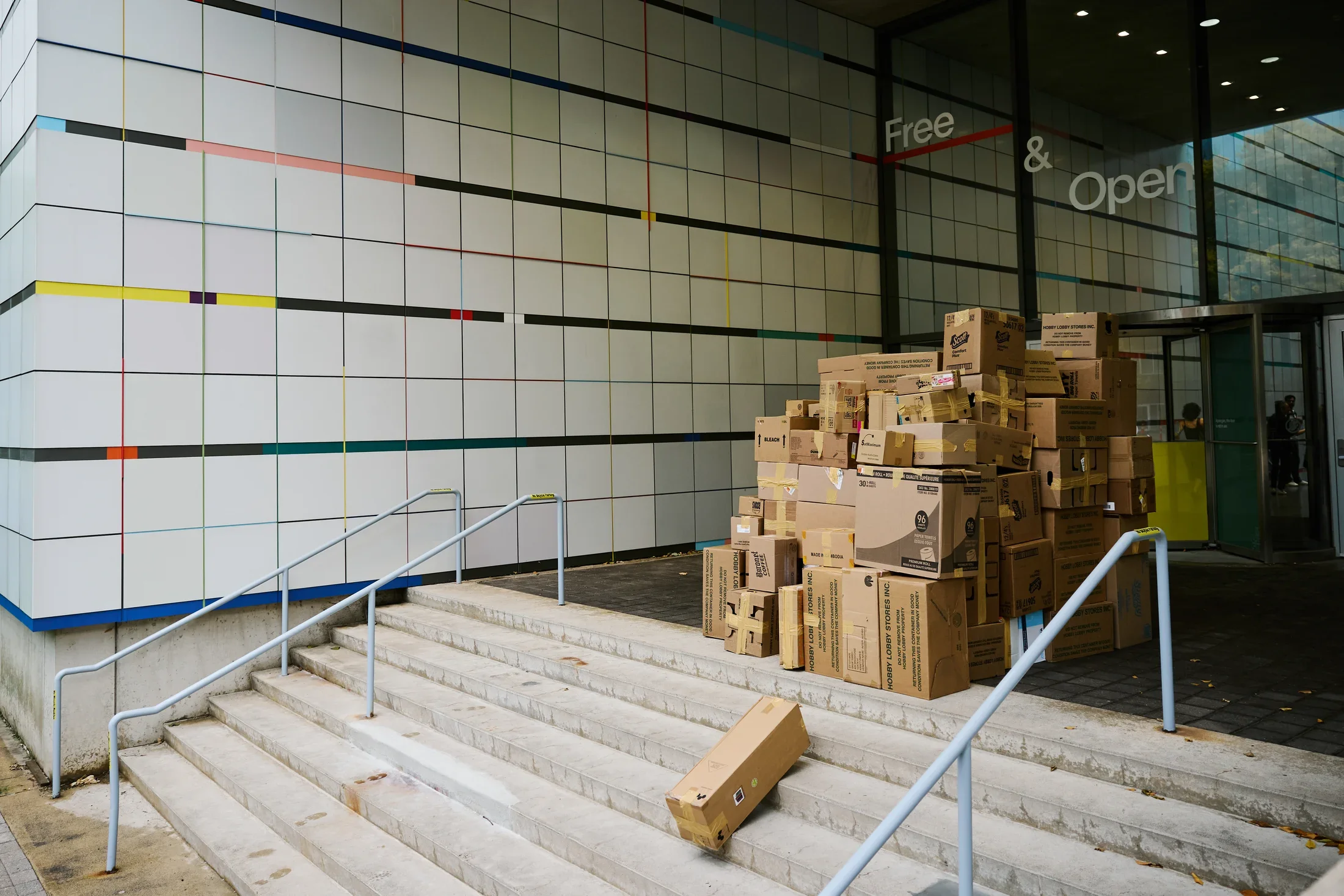 A large pile of cardboard boxes sits next to the glass entrance of a museum. The building's exterior is a grid of white tiles with horizontal lines of colored rectangles. Concrete stairs with light blue handrails lead up to the entrance, with one box resting on a step.
