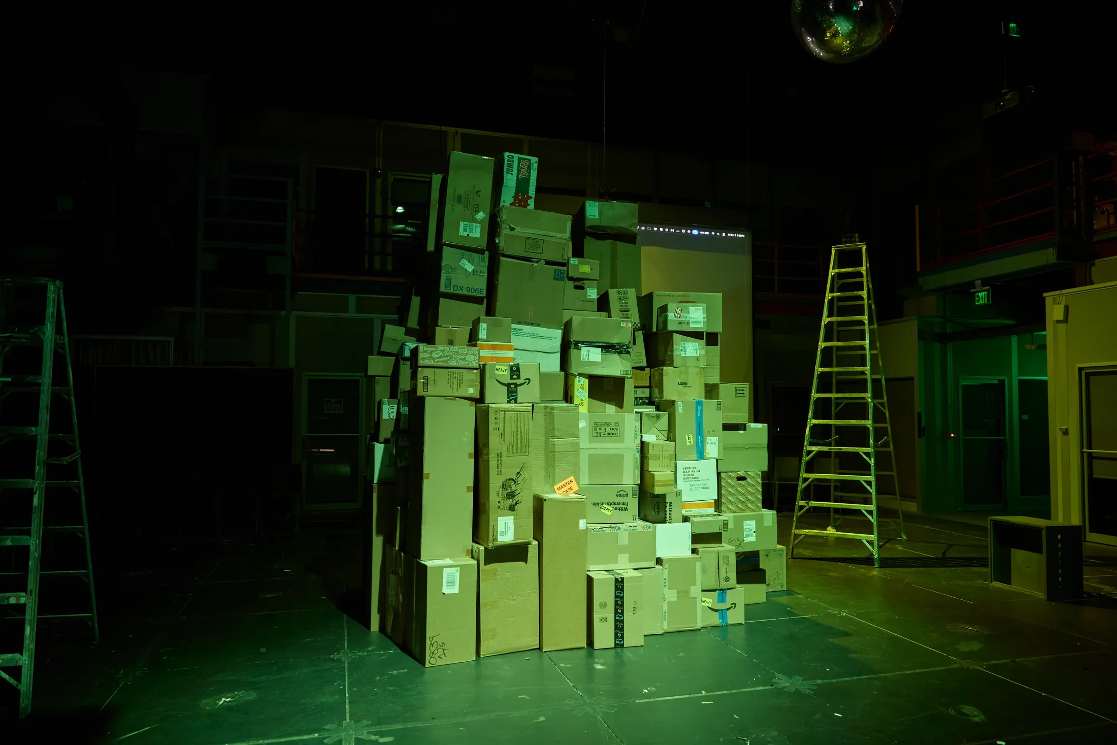 A large, tall pile of cardboard boxes stands in the center of a dark, green-lit room. A tall ladder is on the right, and a disco ball hangs from the ceiling.