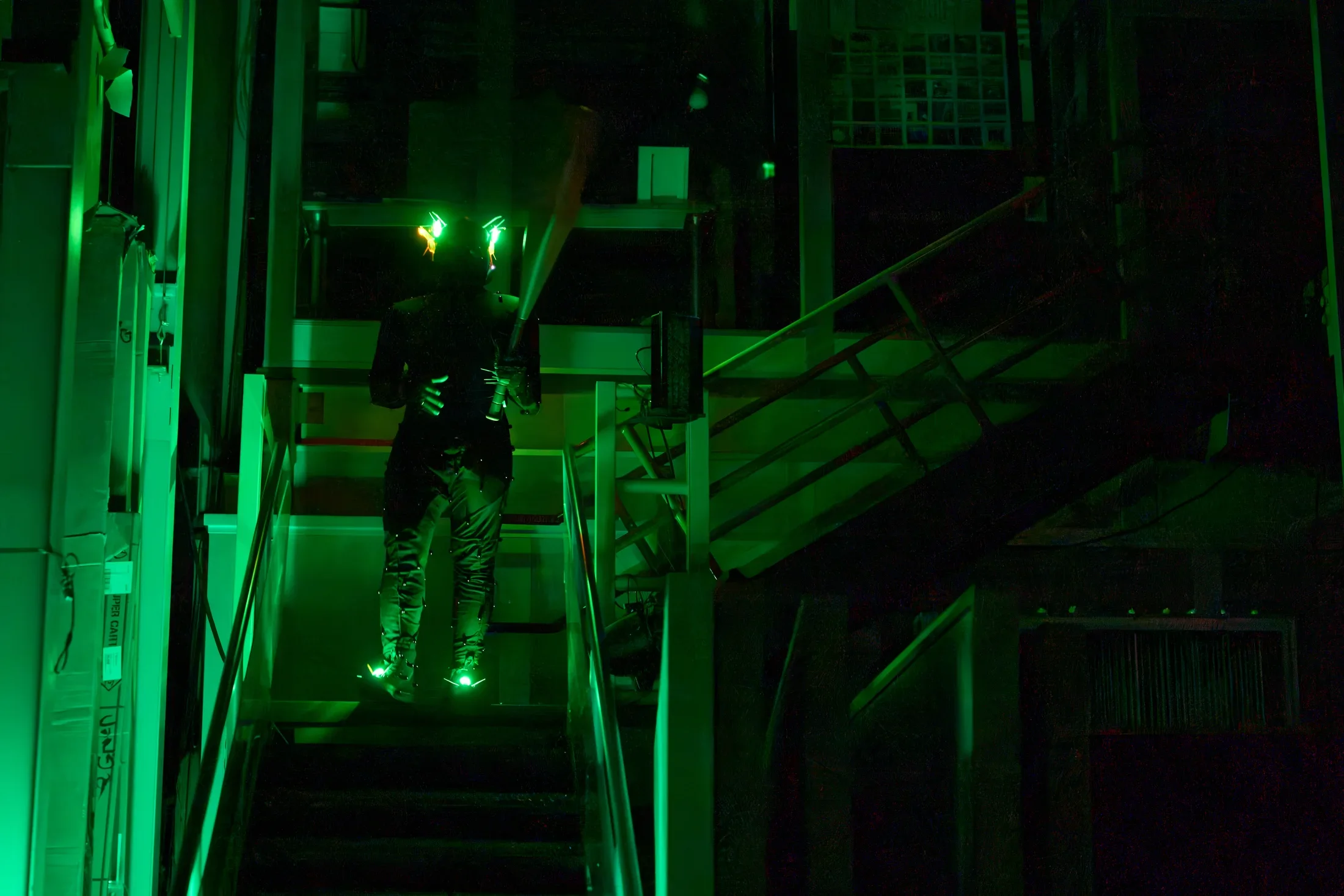 A person in dark clothing stands on a metal staircase with a large baseball bat in hand. Green lights glow from their head and feet, illuminating the dark, industrial setting.