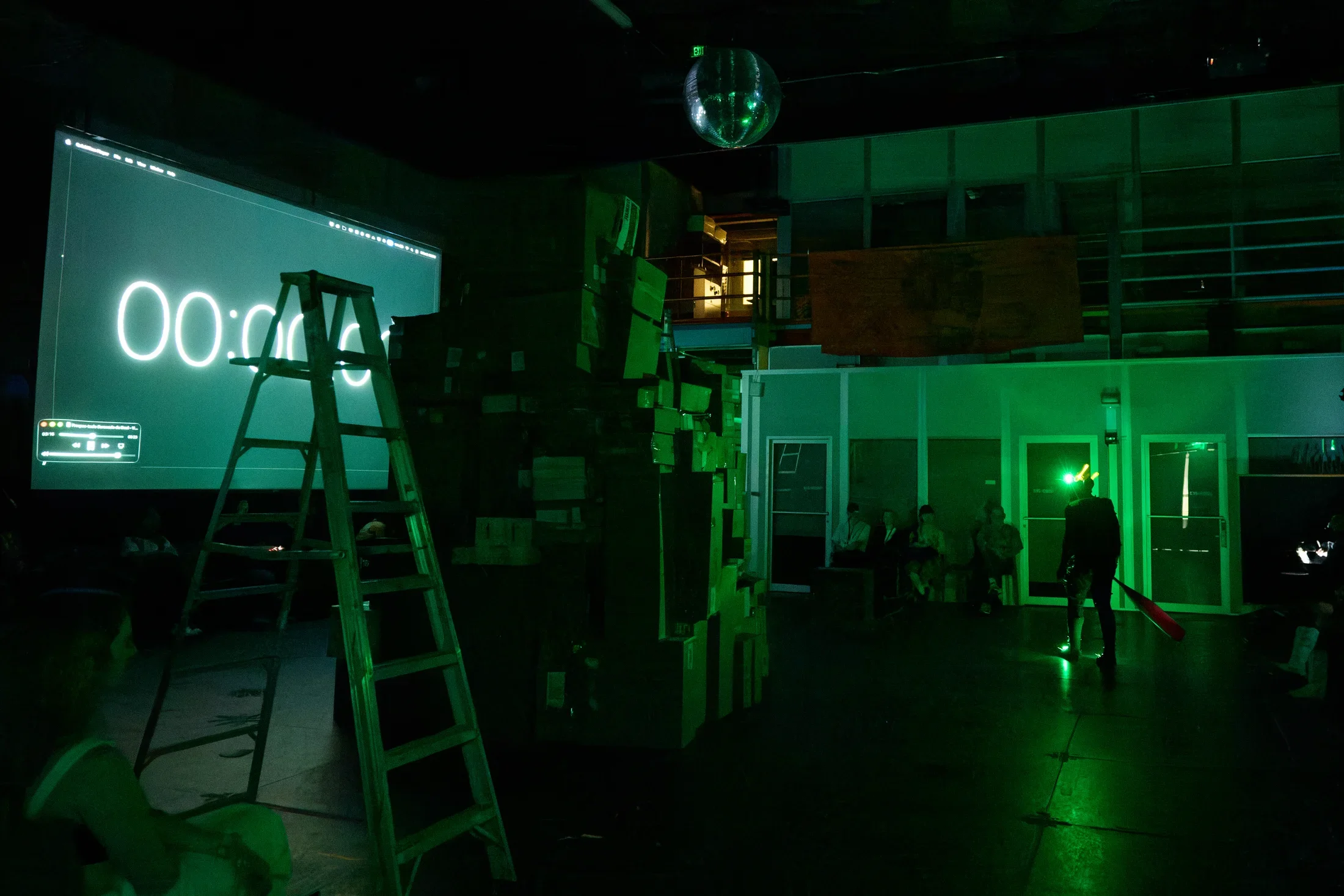 A dark room with green lighting features a large projection screen displaying "00:00" and a wooden A-frame ladder. A tall stack of cardboard boxes stands beside the screen, and a disco ball hangs from the ceiling. Several people are visible, some seated, and one standing with a red baseball bat.