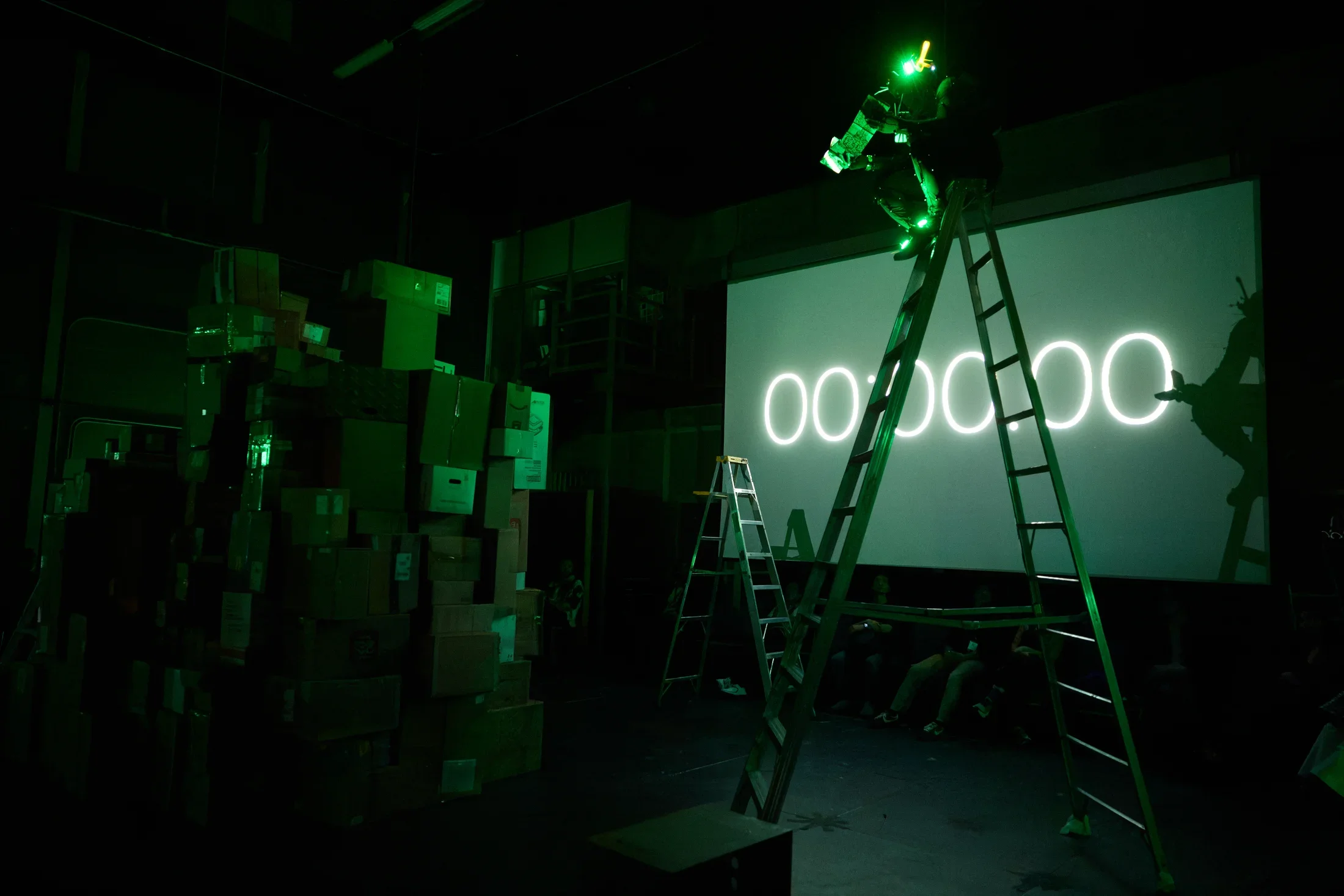 A dark room is illuminated by green light. A person on a tall ladder aims a green laser at a large screen displaying "00:00:00". To the left, a tall stack of cardboard boxes is visible, and several people sit on the floor in front of the screen.