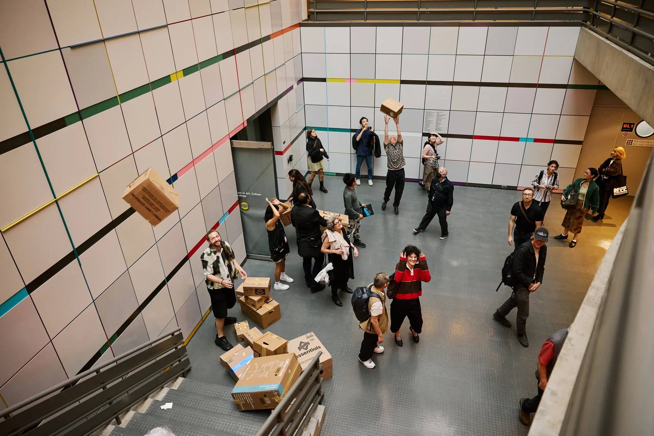 A group of people interact with cardboard boxes in a multi-level modern building. Some boxes are stacked on stairs, others are held, and one is in mid-air. The white tiled walls are decorated with horizontal colored stripes.
