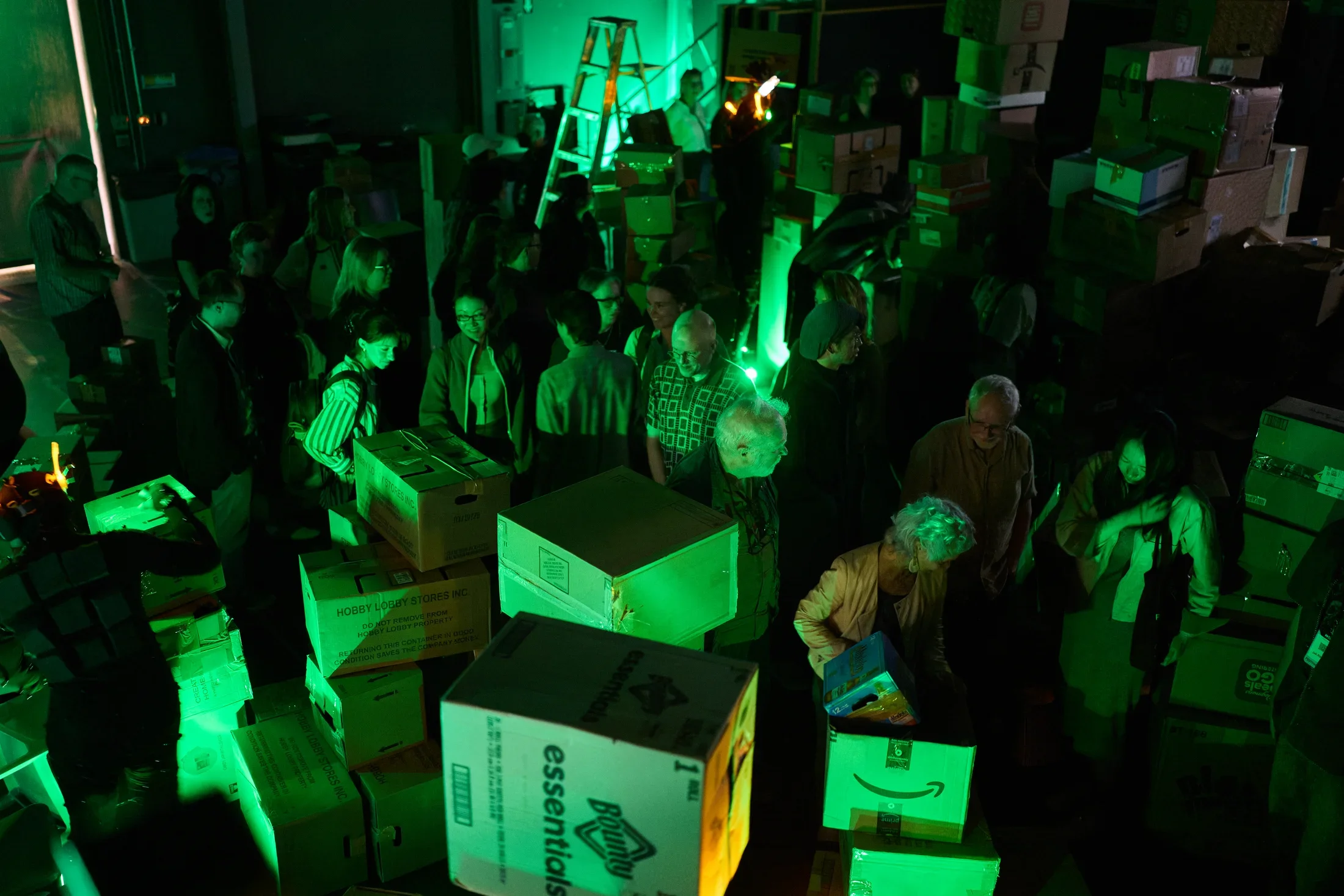 A large group of people stands amidst numerous cardboard boxes in a dimly lit room, illuminated by a strong green light. Boxes of various sizes are stacked and scattered throughout the space.
