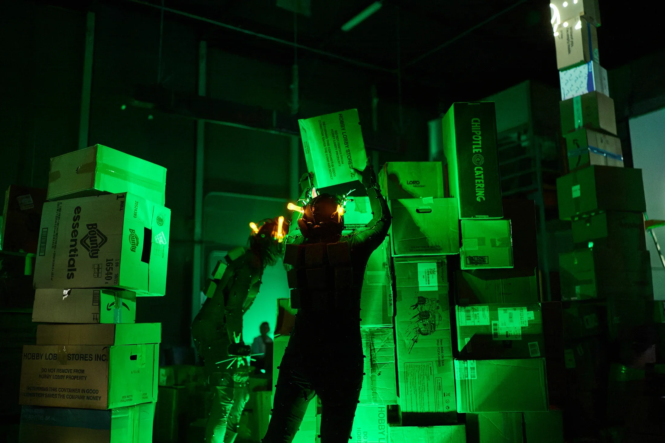 Two figures in dark, padded costumes with glowing orange headpieces interact with stacks of cardboard boxes under green light. One figure holds a large box above their head, while the other leans over a pile of boxes.