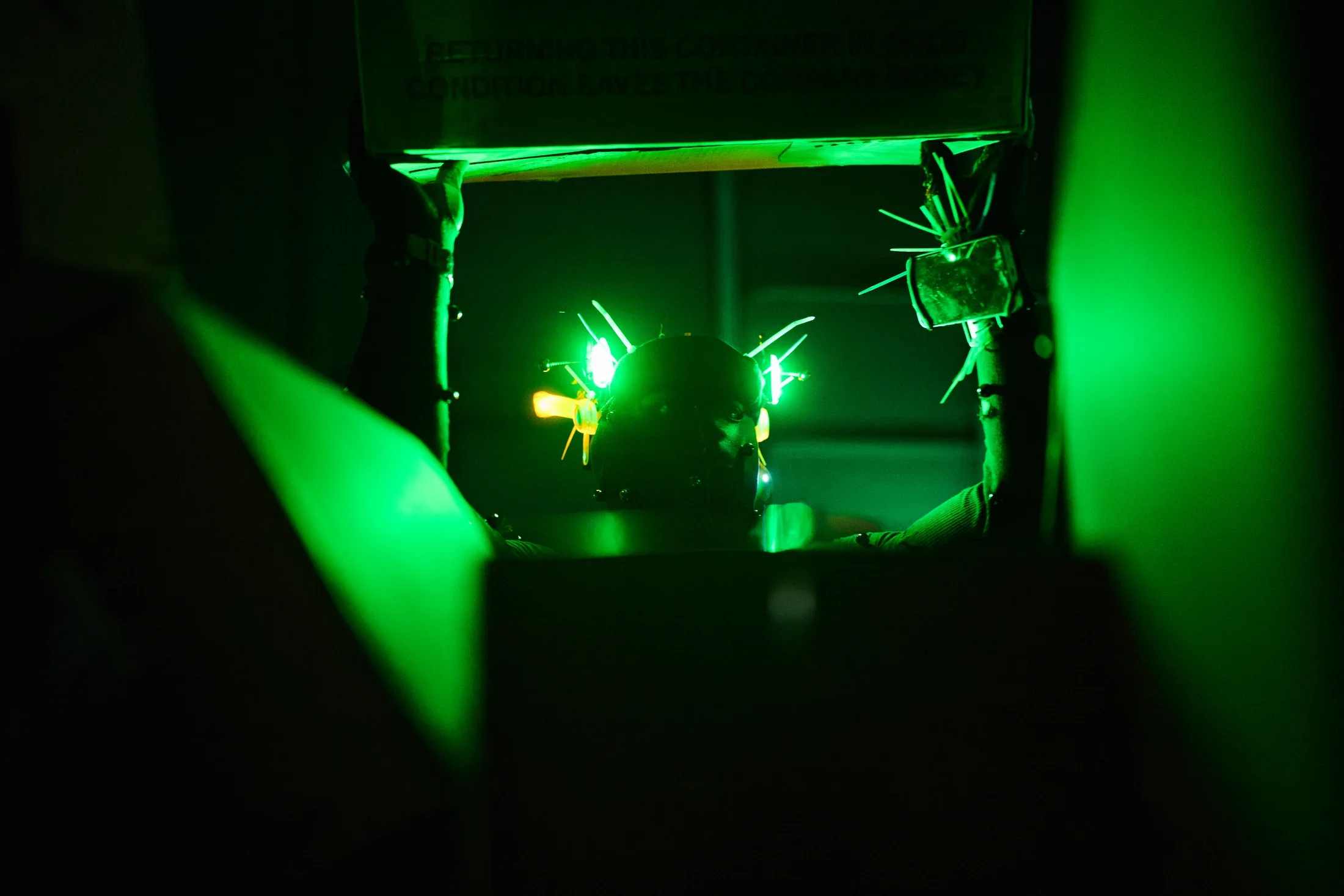 A person's head, wearing a dark helmet with bright green spiky lights, is illuminated by green light within a dark space. The person holds up a box while surrounded by other cardboard boxes.