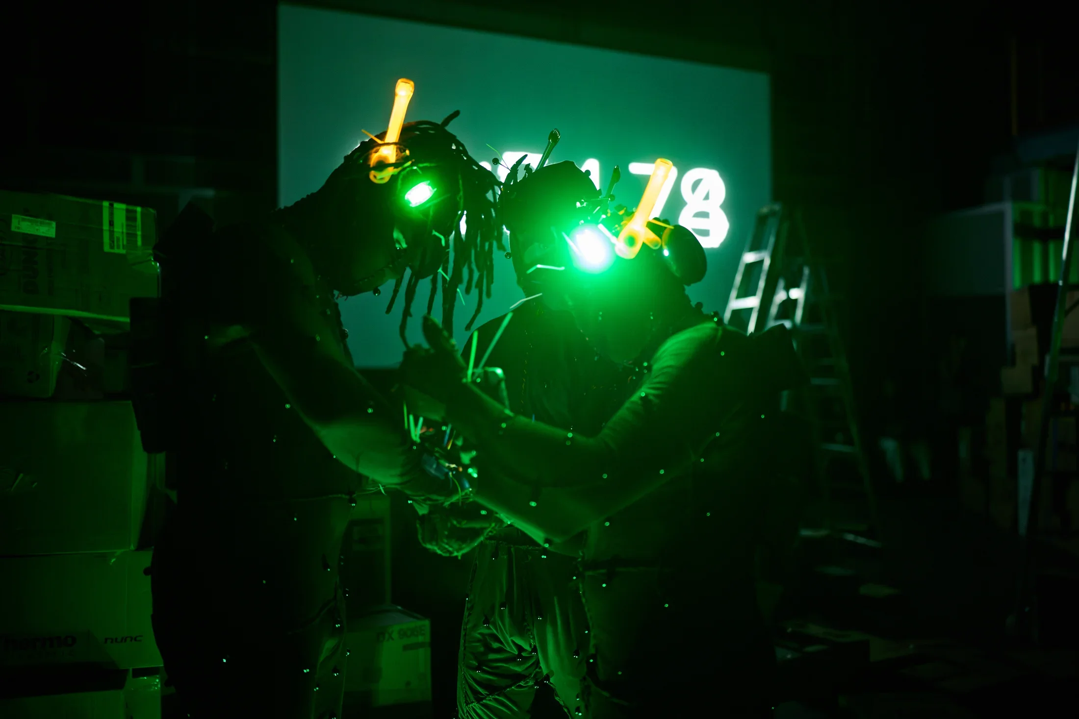 Three figures in dark, motion-capture-like suits with glowing green headgear stand facing each other in a dimly lit space. Small pearl-like dots are visible on their bodies, and the number 78 is projected in green light on a screen behind them.