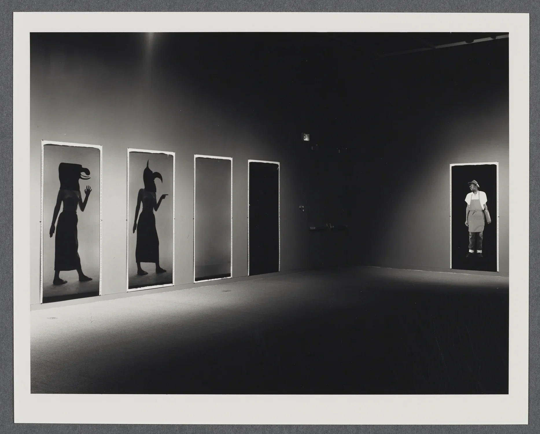 Black and white image of large polaroids lining the gallery wall. Some polaroids are left blank while others have figures.
