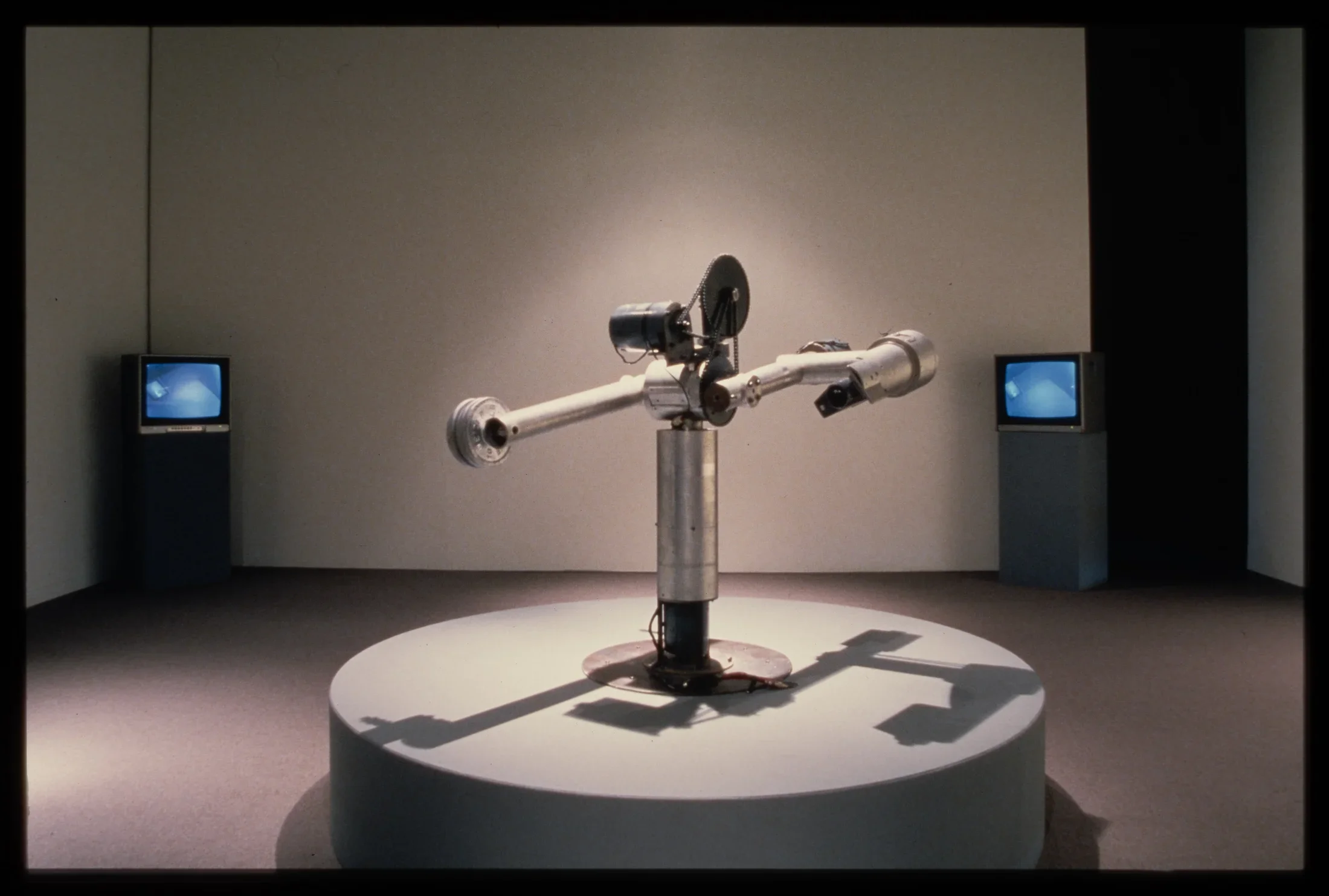 A mechanism sits atop a circular pedestal. A silver cylinder acts as a base while two cylindrical arms are attached by gears.