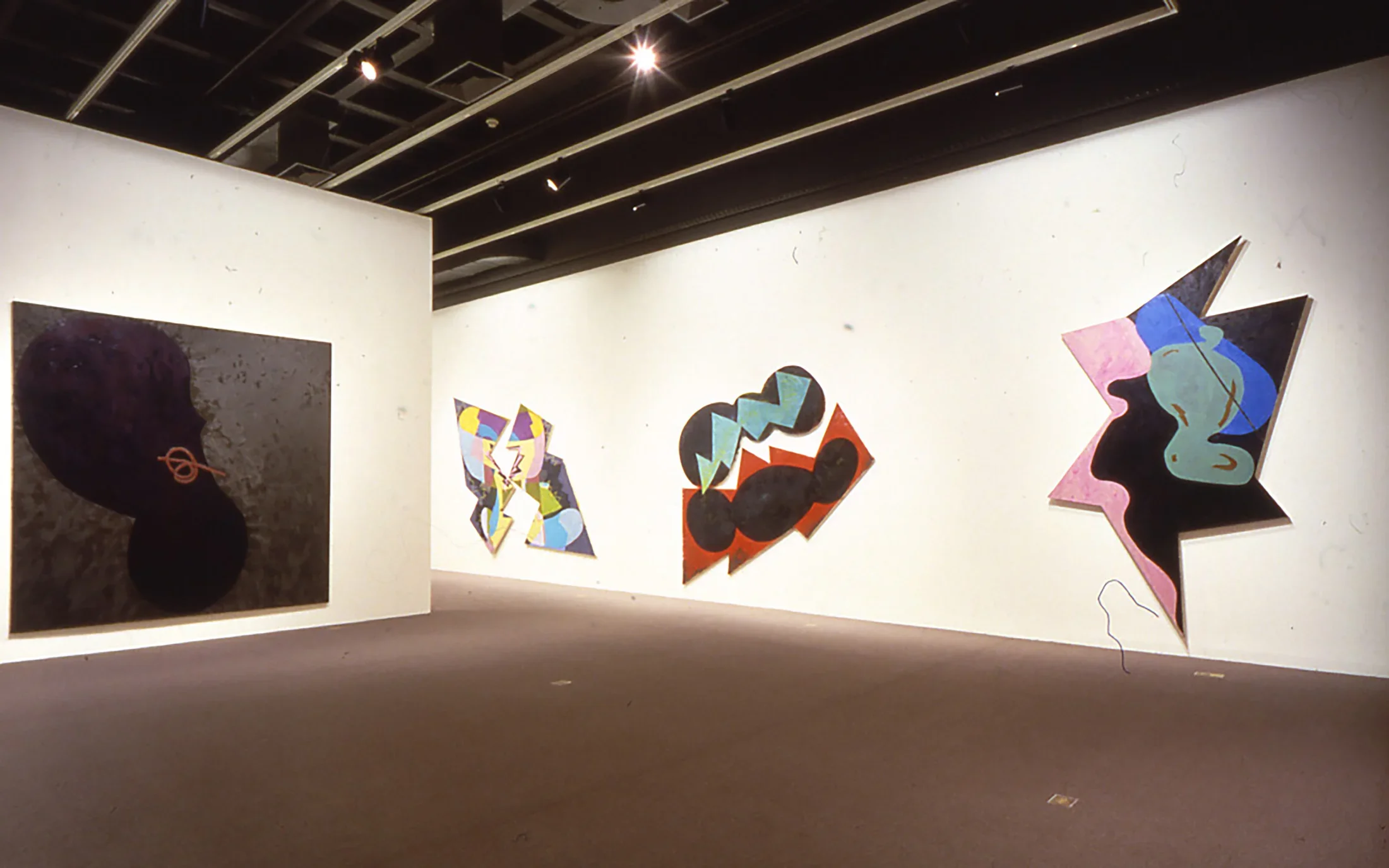A large dark rectangle shaped painting is hung on one wall and 3 vivid abstract multi shaped paintings are hung on another.