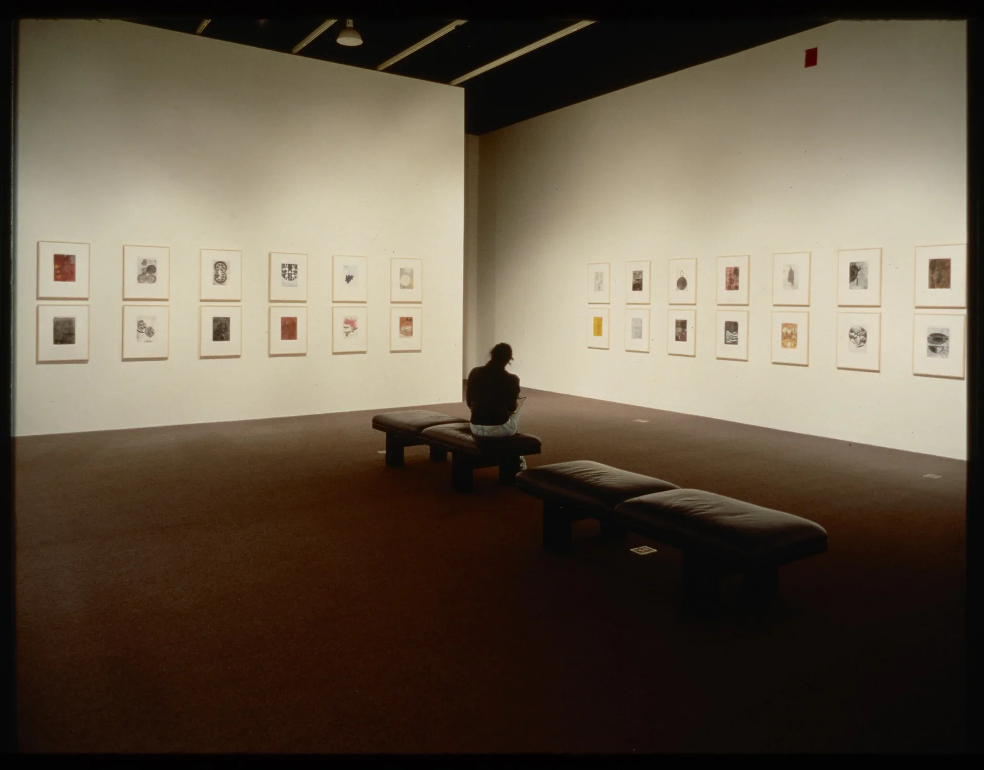 A visitor sits on gallery benches while viewing the framed works that are hung on the walls around them.  