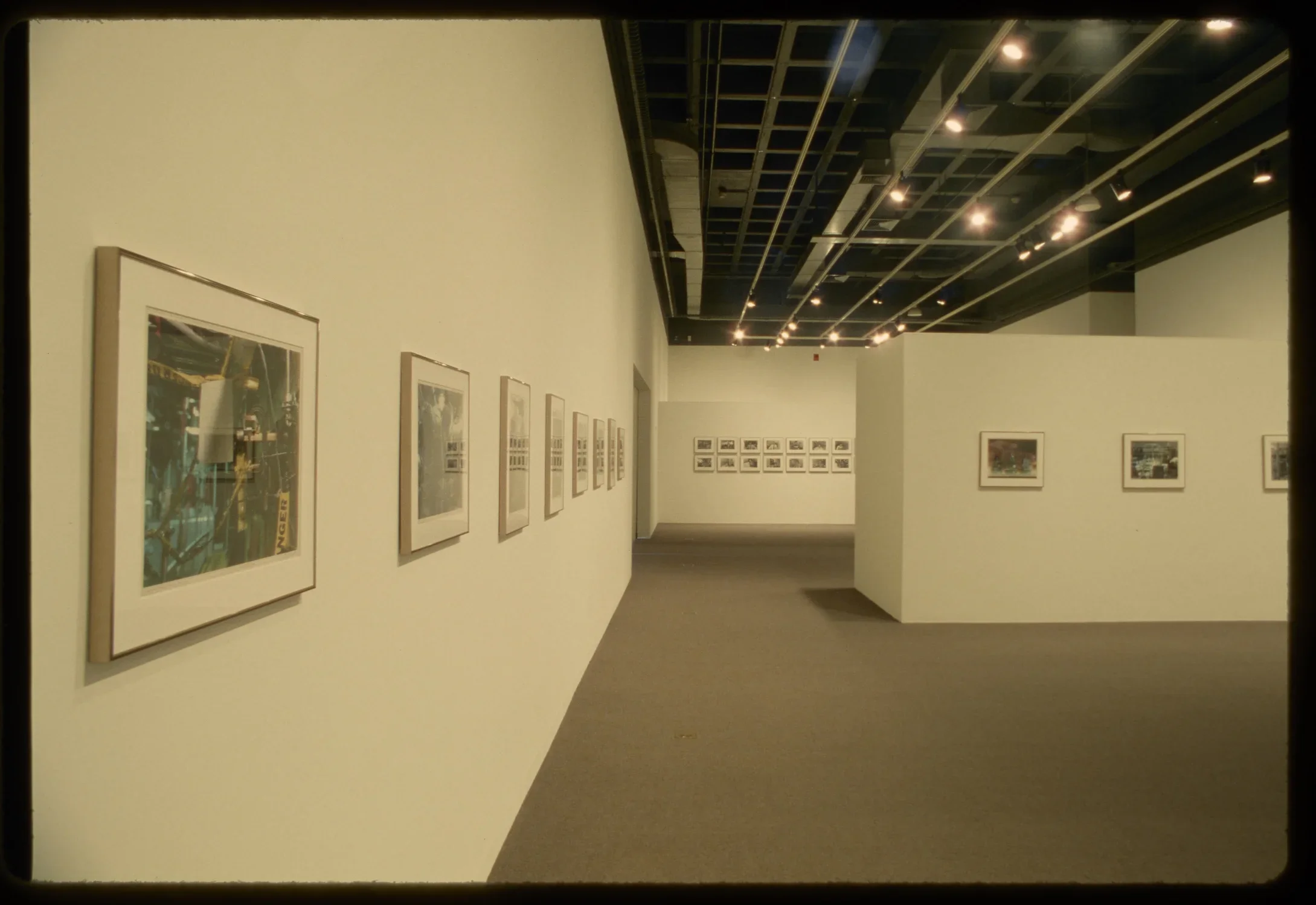 Eleven small framed photographs hang in the middle of gallery walls. Fourteen small photographs hang in the distance.