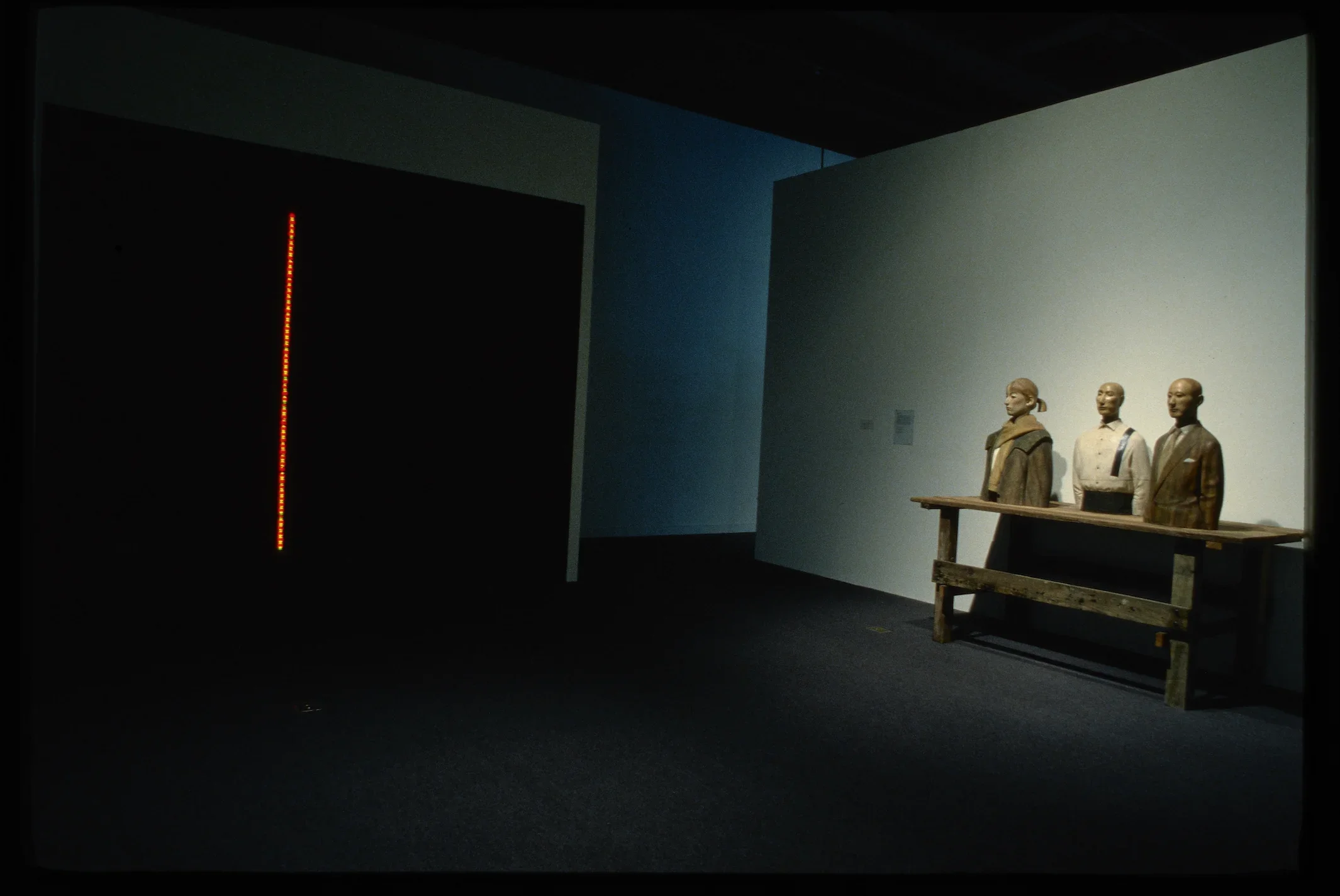 A tall strip of orange light stands out in a dark room, on the right is a sculpture of 3 people from the waist up atop a table.