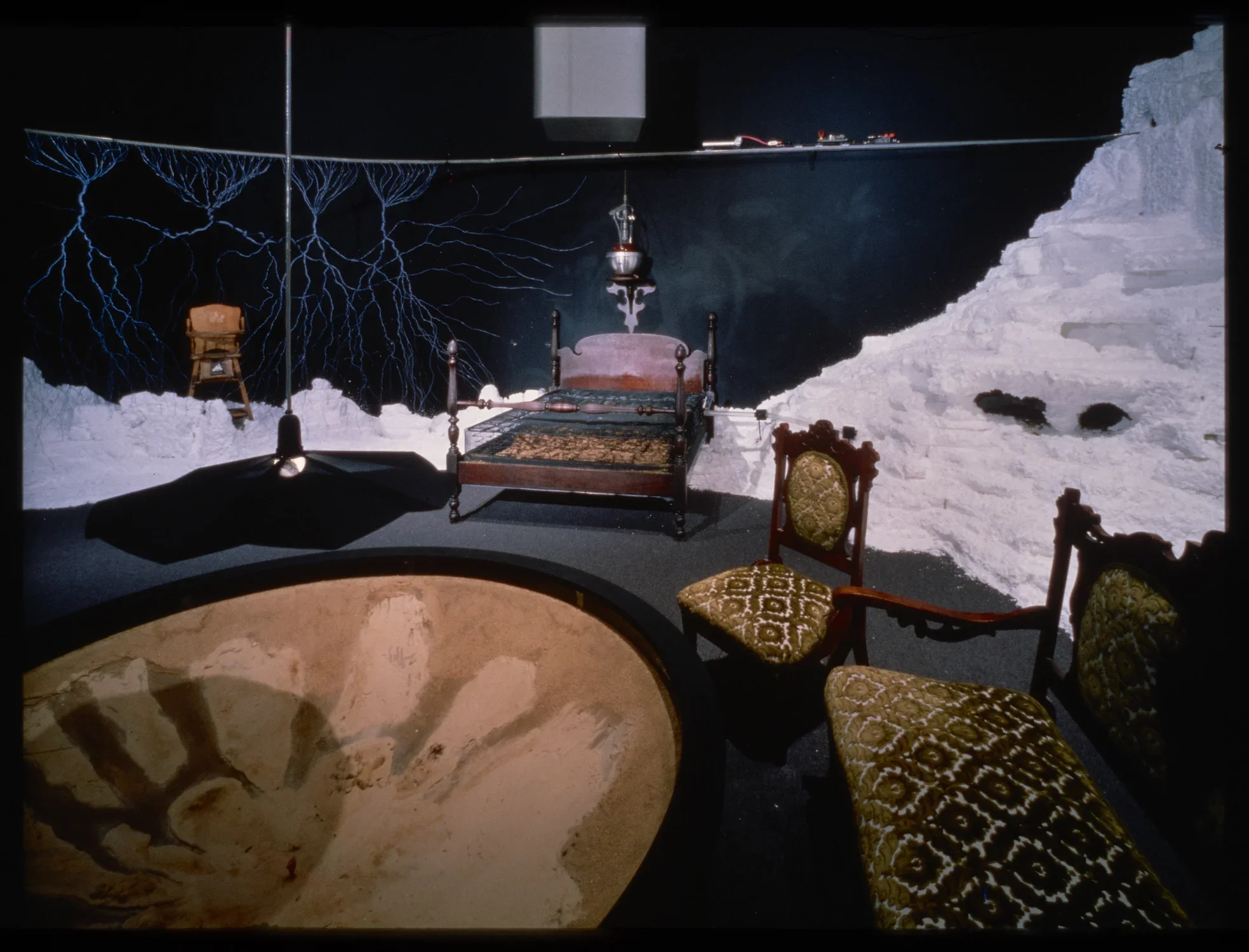 An installation of a room with a table, chairs, and bed. These elements sit before a constructed white mountain.
