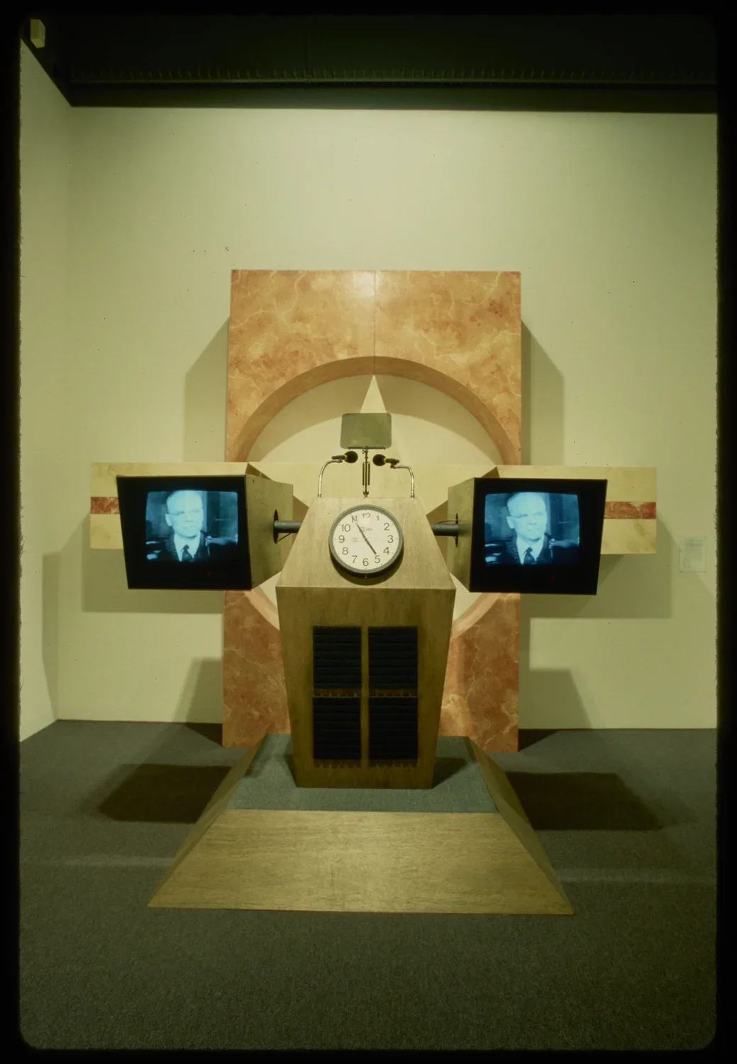 A podium has a clock with two monitors on each side showing a television broadcast.