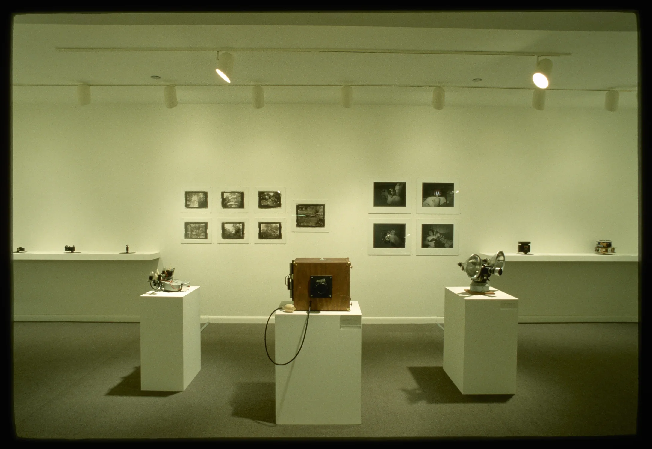 Different types of cameras sit on pedestals and on shelves. Black and white photographs are pinned to the gallery wall.