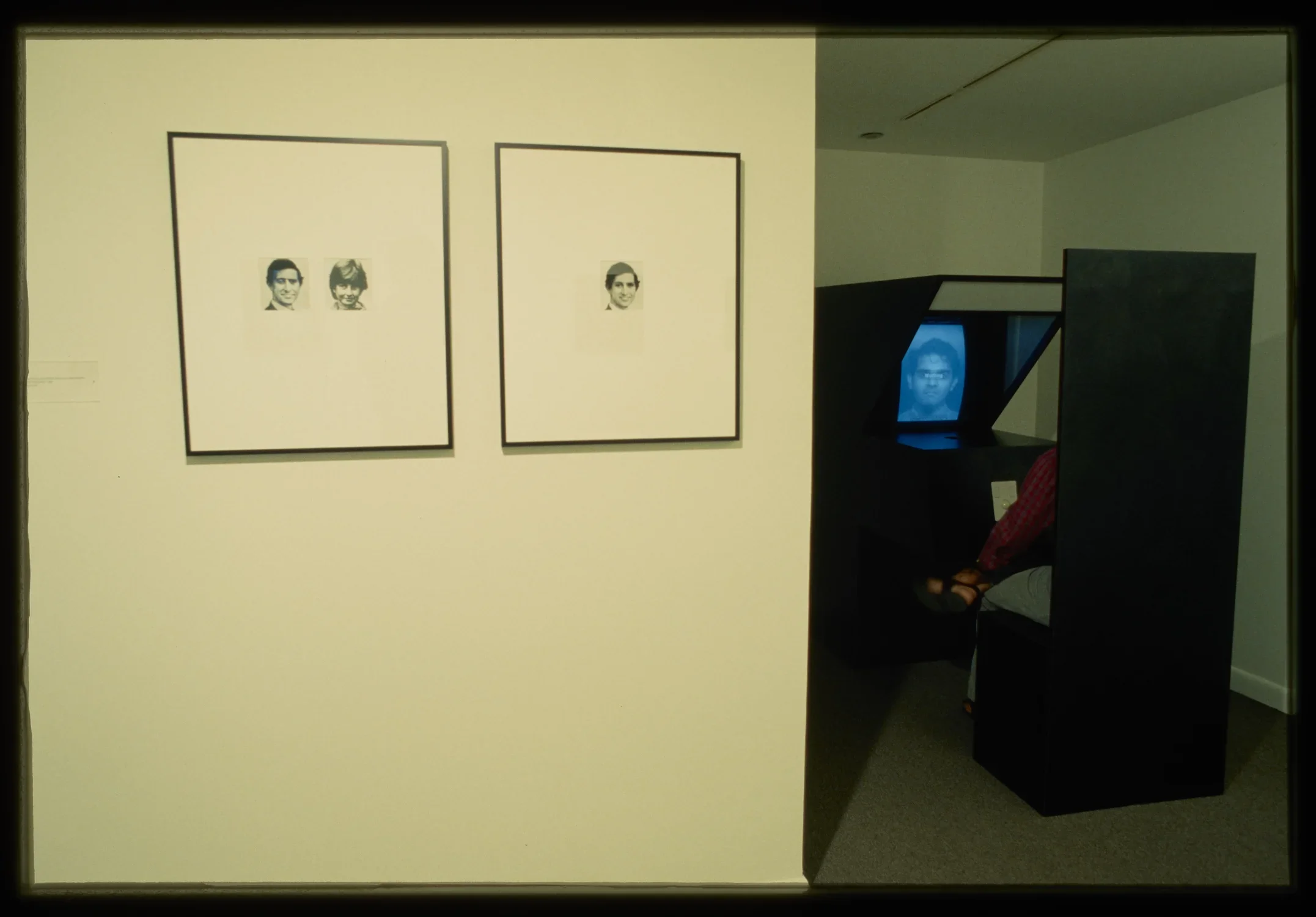 Two images hang on the gallery wall while a photobooth is set up in a dark corner of the gallery. 
